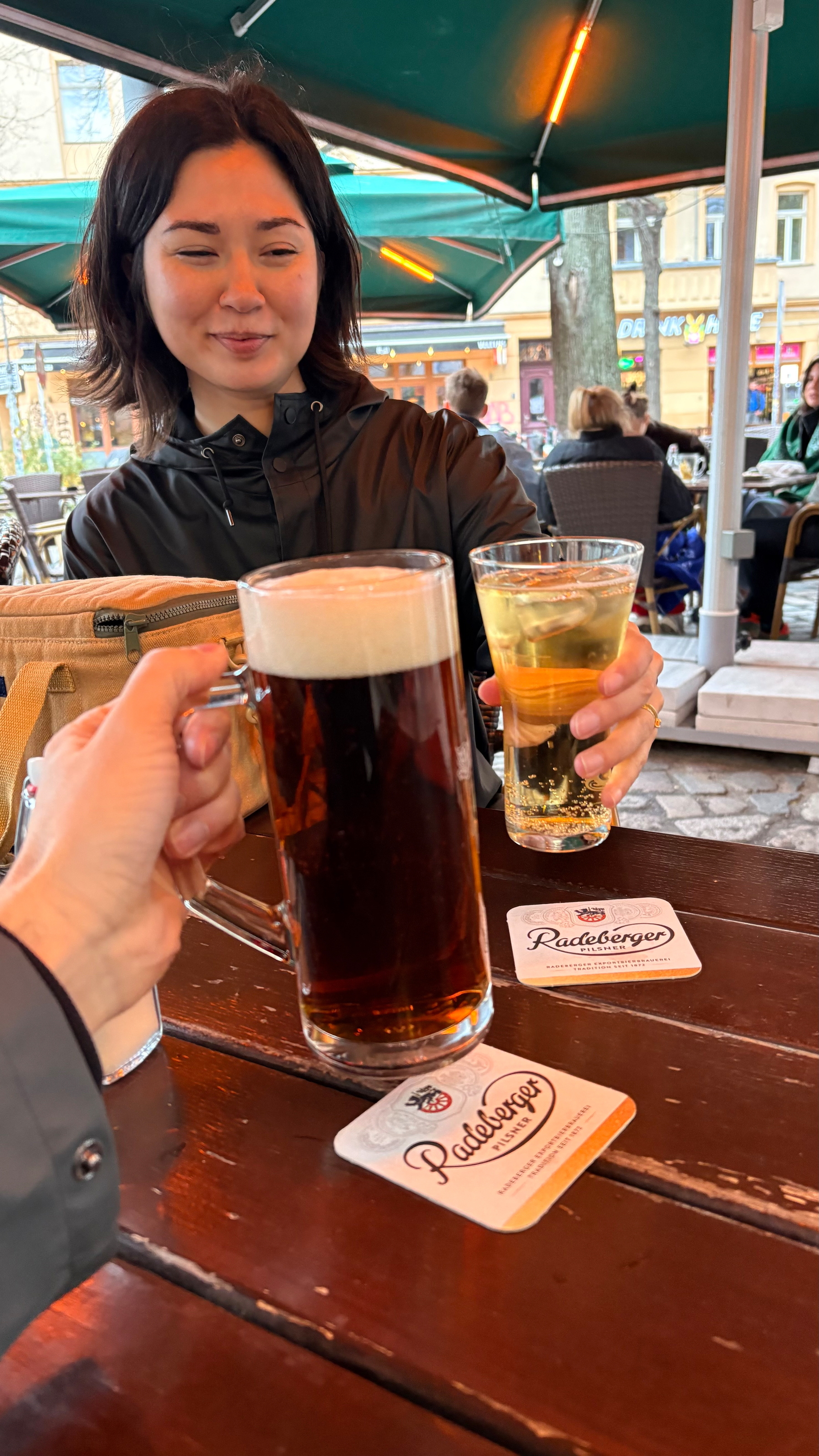 Auto-generated description: Two people are toasting with drinks at an outdoor wooden table under an umbrella.