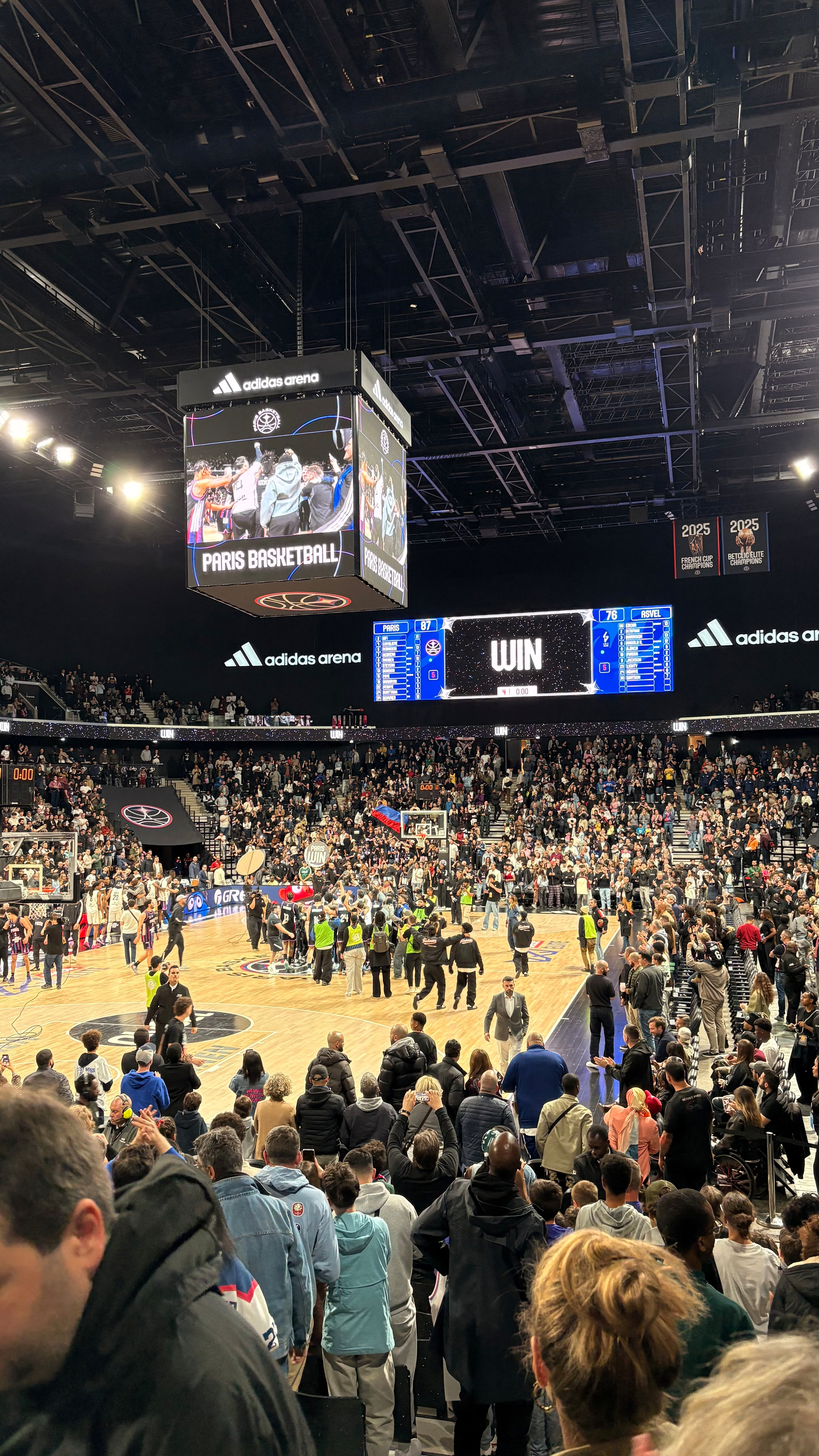 Crowd celebrating on a basketball court with a scoreboard displaying WIN at Adidas Arena.