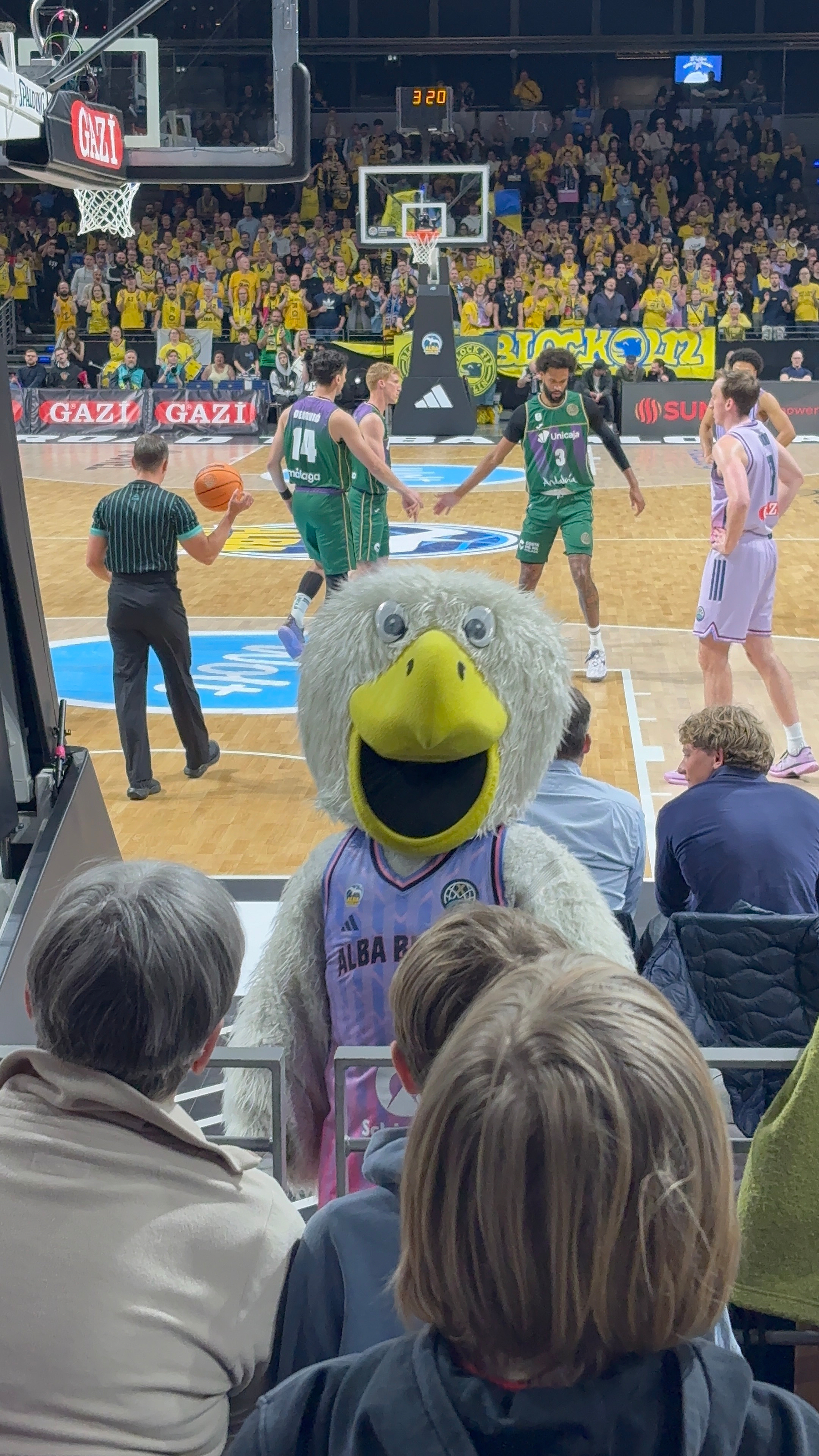 Auto-generated description: A large, plush eagle mascot stands in the foreground while a basketball game is actively taking place in the background.