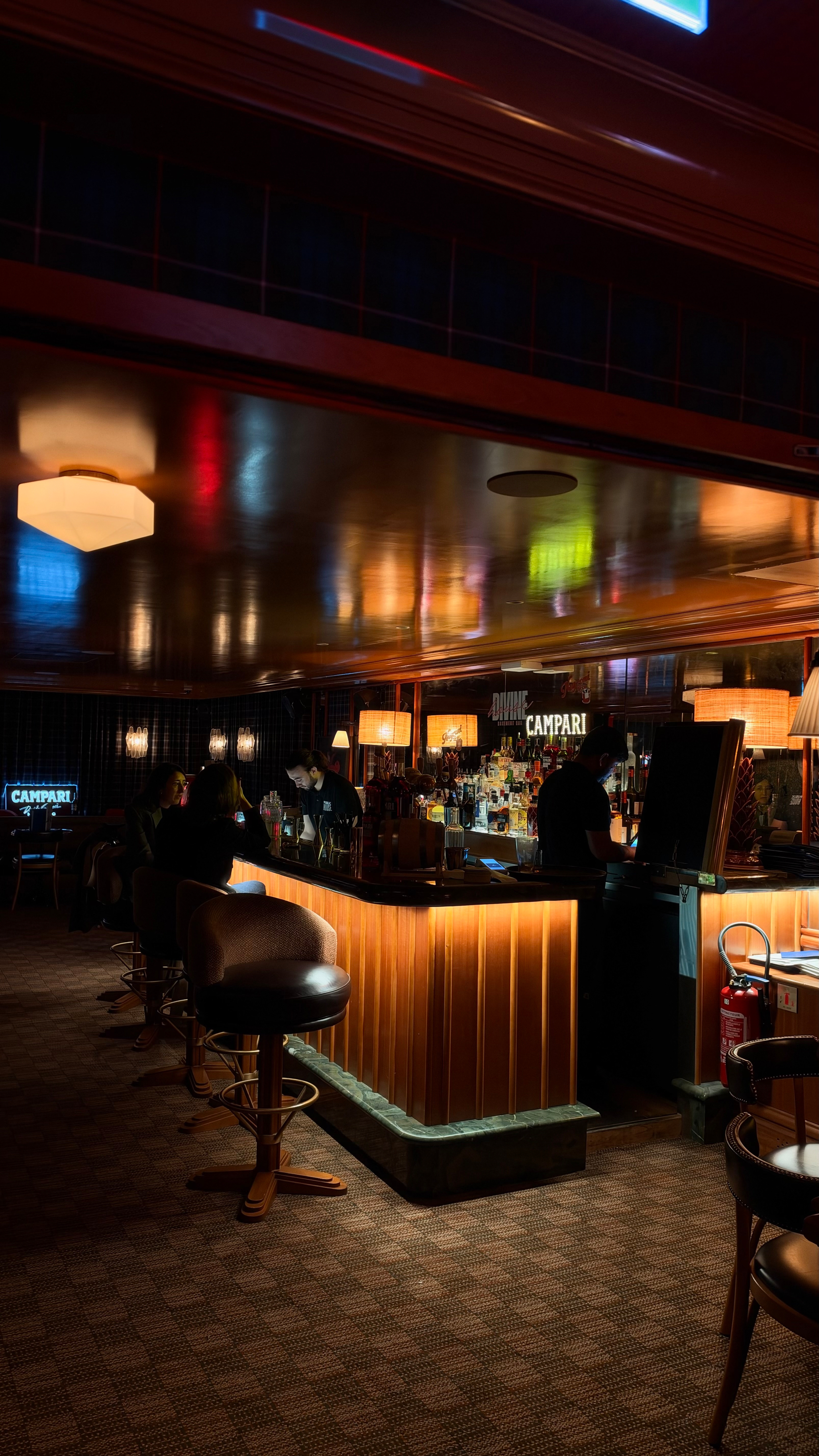 A dimly lit bar with illuminated counters, bar stools, and a few patrons seated and interacting with the bartender.