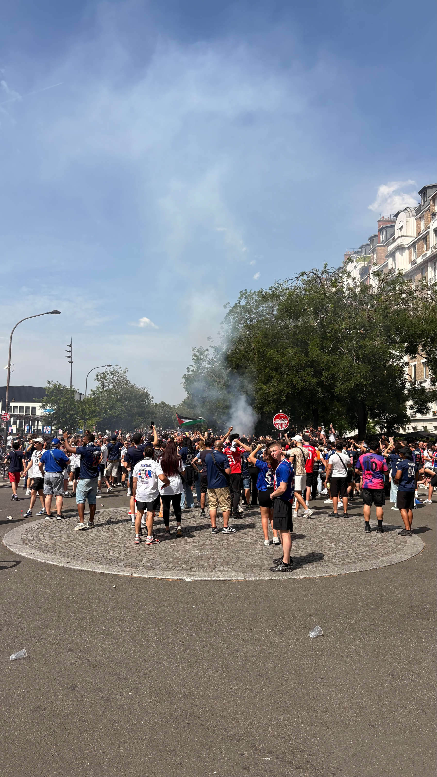 A large group of people gathers around a central area on a city street, with some smoke rising into the sky.