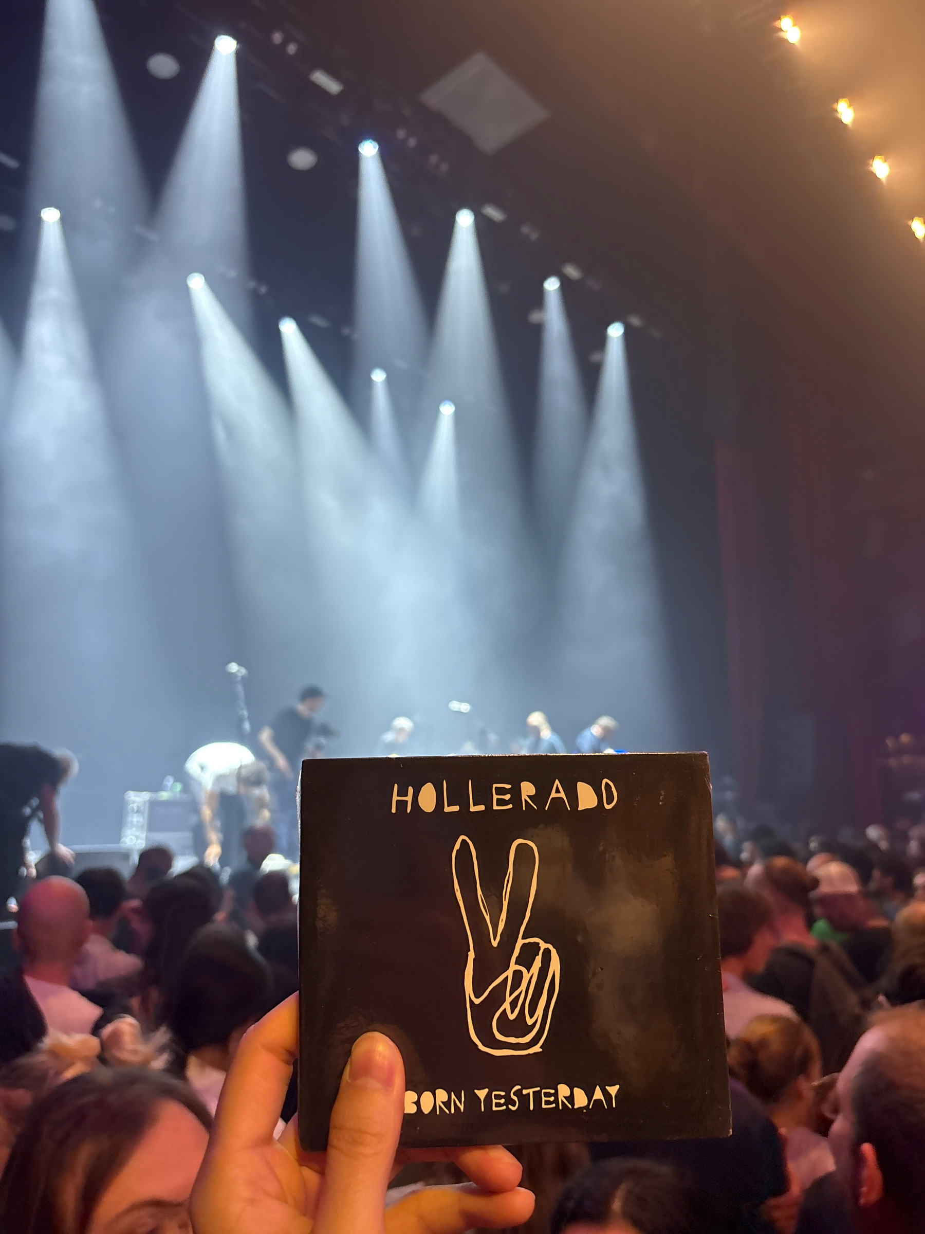 Auto-generated description: A hand holds a Hollerado Born Yesterday album cover in front of a crowded concert stage with bright spotlights and band members.