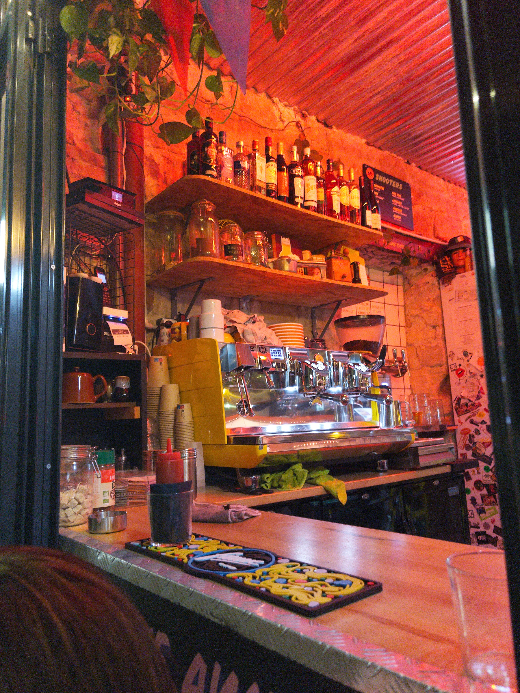 Auto-generated description: A cozy café counter is adorned with a variety of coffee-making equipment, shelves of bottles, and decorative plants.