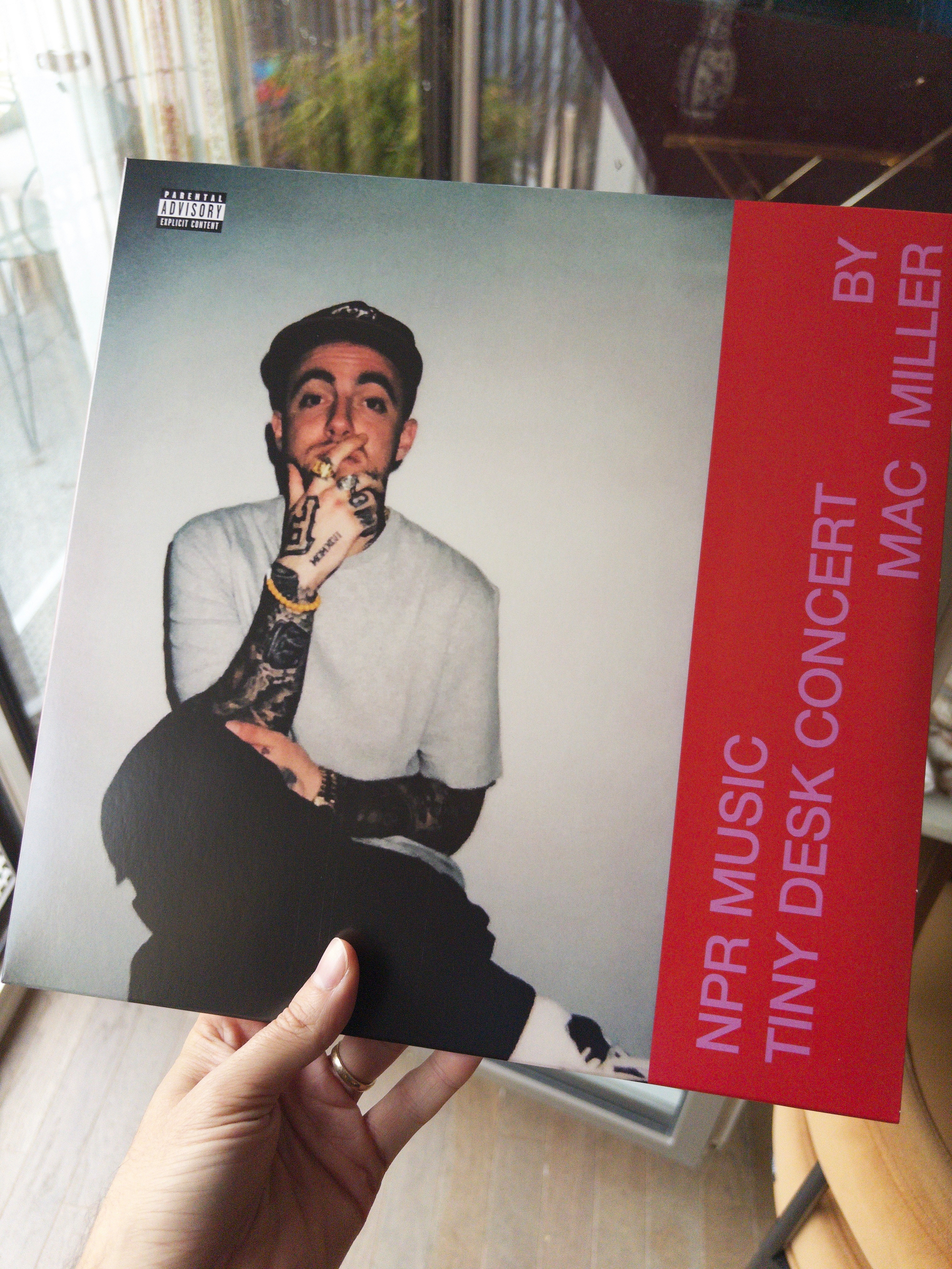 A person is holding a vinyl record cover featuring a photo of Mac Miller with text that reads NPR Music Tiny Desk Concert by Mac Miller and a parental advisory label.