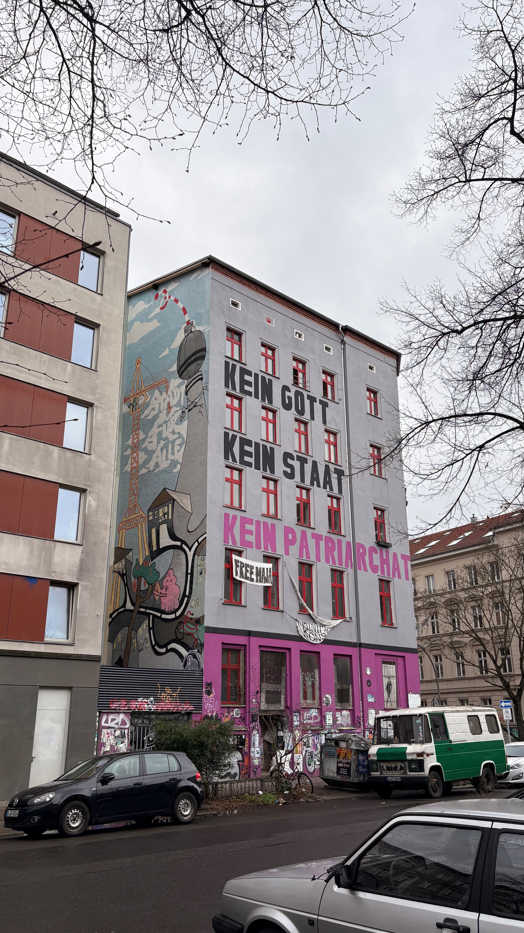 Auto-generated description: A multi-story building with vibrant graffiti art and political slogans is shown surrounded by parked cars on a street.