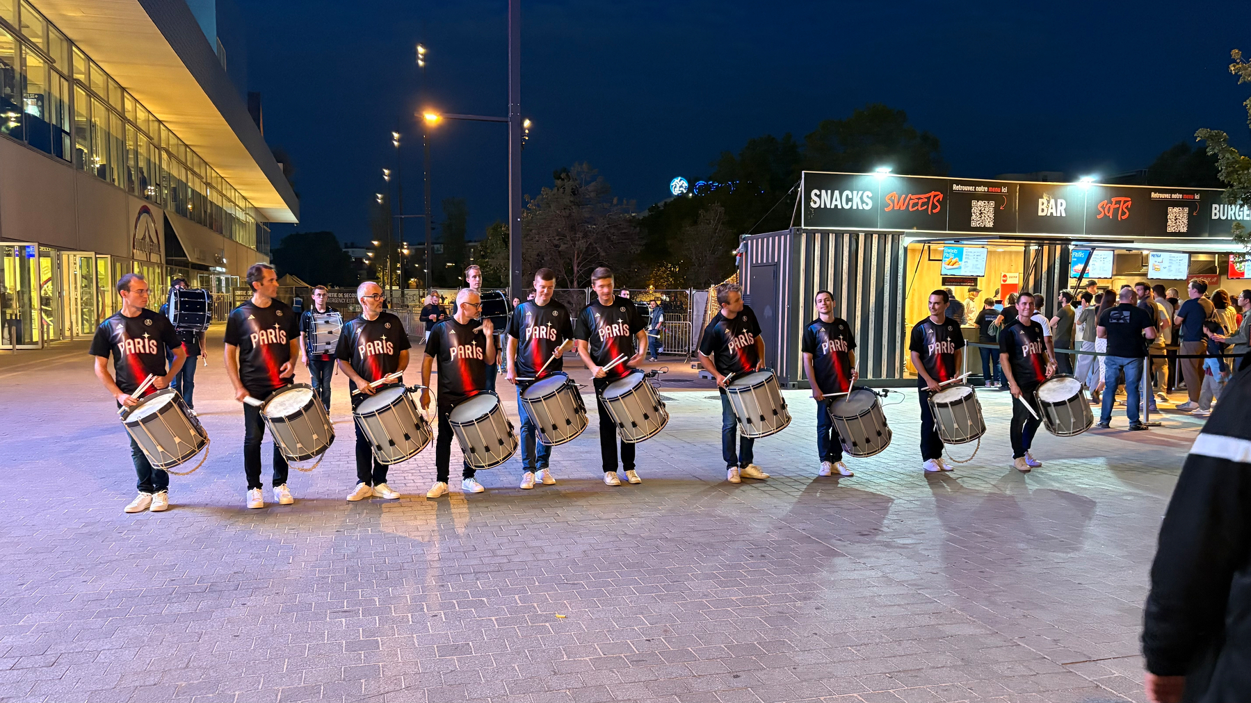 A line of drummers wearing matching black and red Paris shirts perform outdoors at night near a snack and bar stand.