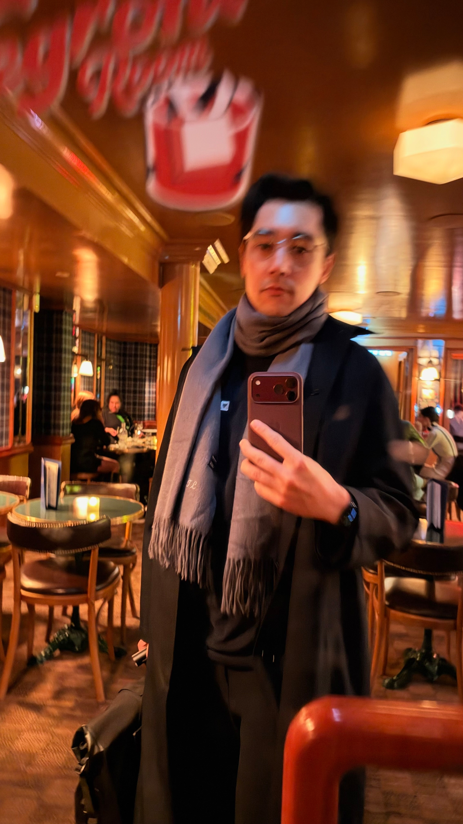 A person wearing glasses and a scarf takes a selfie in a warmly lit, cozy restaurant or bar with wooden furniture and other patrons in the background.