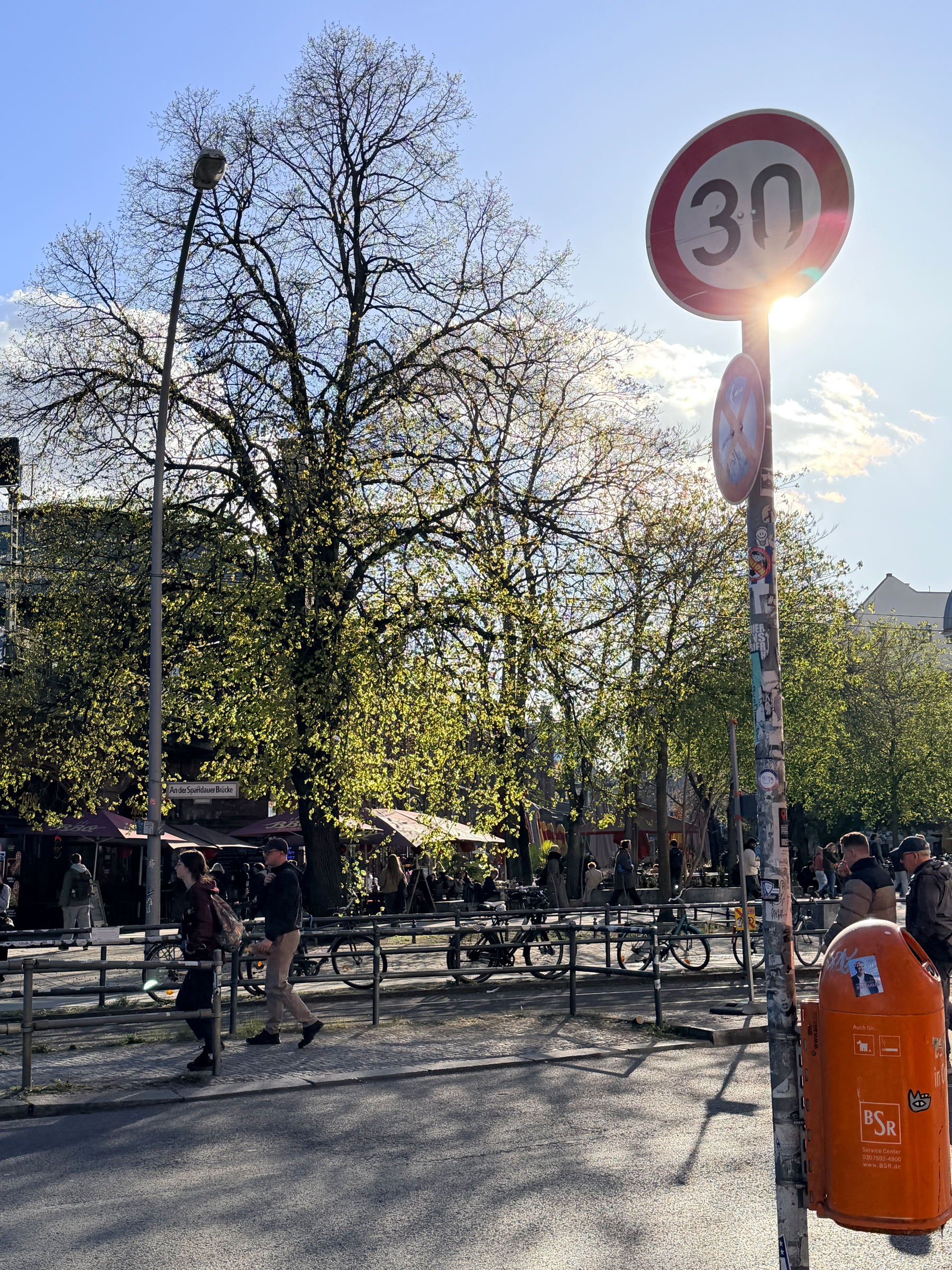 Auto-generated description: A street scene featuring a 30 km/h speed limit sign, people walking and cycling, and trees with bright sunlight filtering through the leaves.