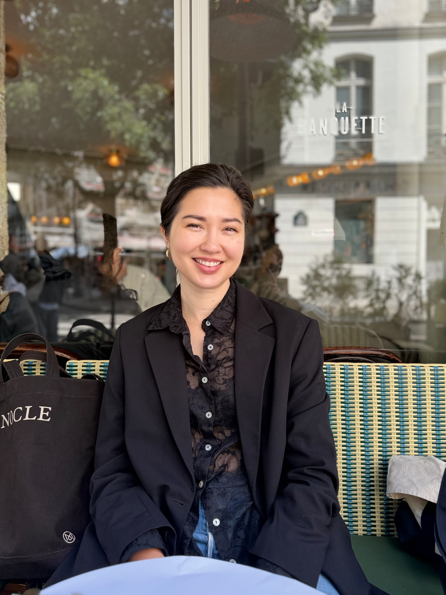 Auto-generated description: A person is sitting and smiling at an outdoor café with a bag beside them.