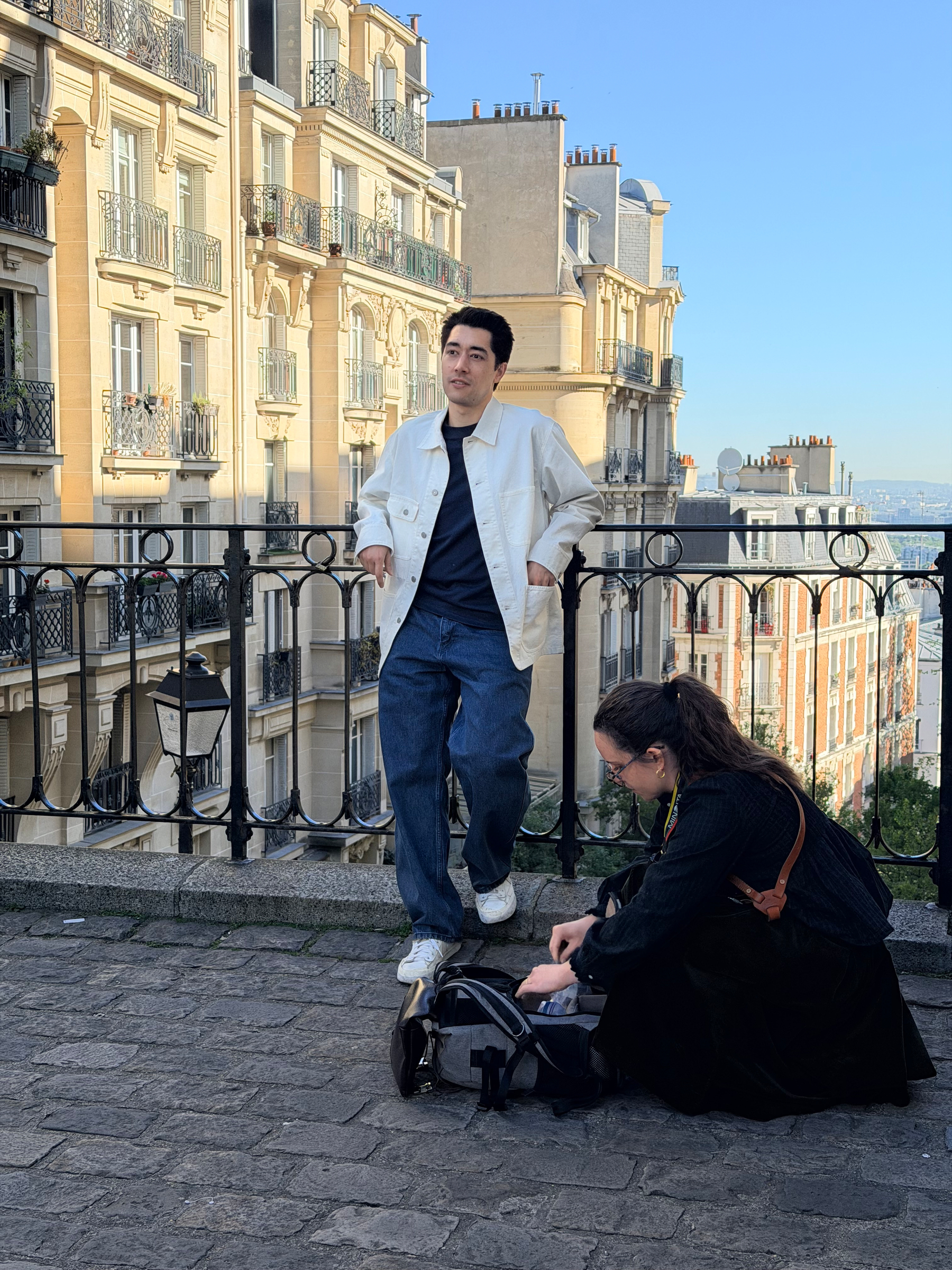 Auto-generated description: A man is leaning against a railing, while a woman is crouching near a bag in front of a row of classic buildings.