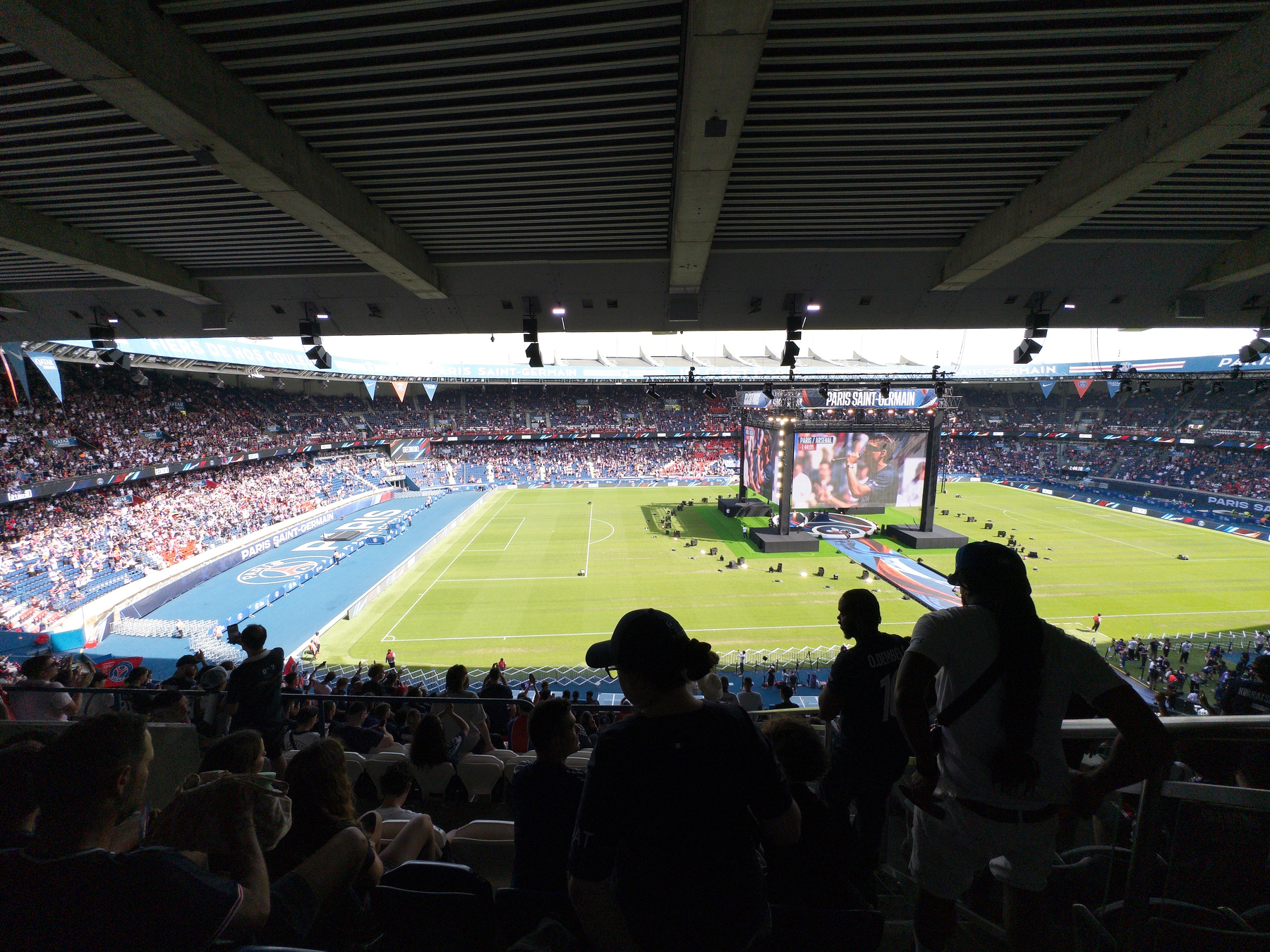 A crowded stadium with a large screen is set for a sporting event, featuring a green soccer field surrounded by stands filled with spectators.