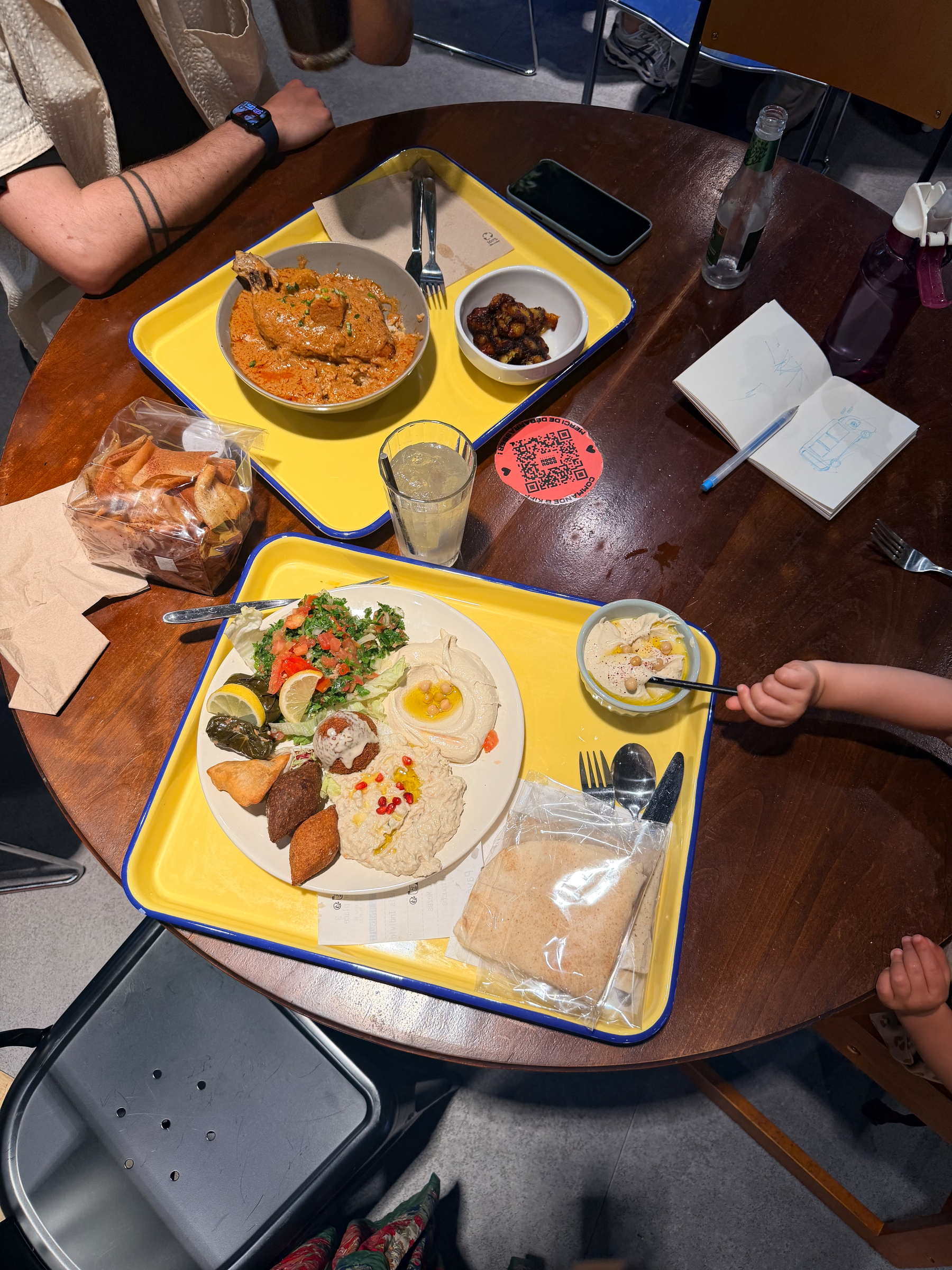 A table with various dishes including a curry, a Mediterranean platter, and a child's hand reaching for food, surrounded by drinks, bread, notebooks, and napkins.
