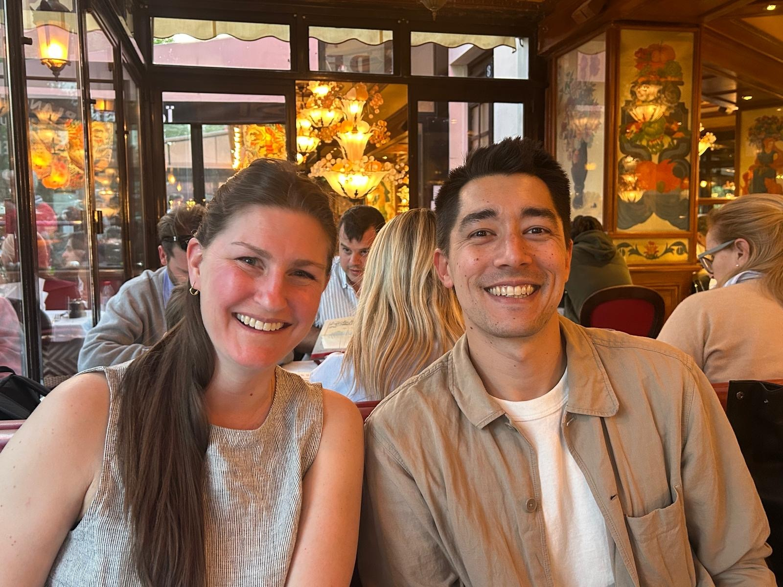 Two people are sitting and smiling in a warmly lit restaurant with other diners in the background.