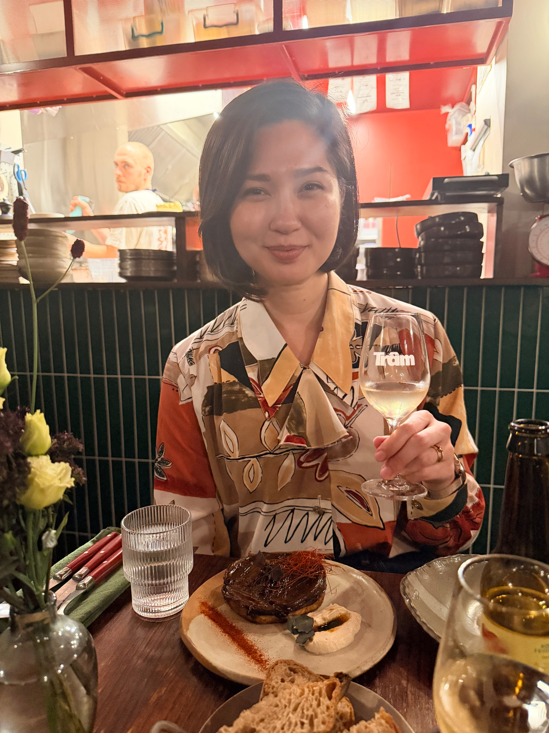 A person is sitting at a table in a restaurant, holding a glass of white wine, with plates of food and bread in front of them.