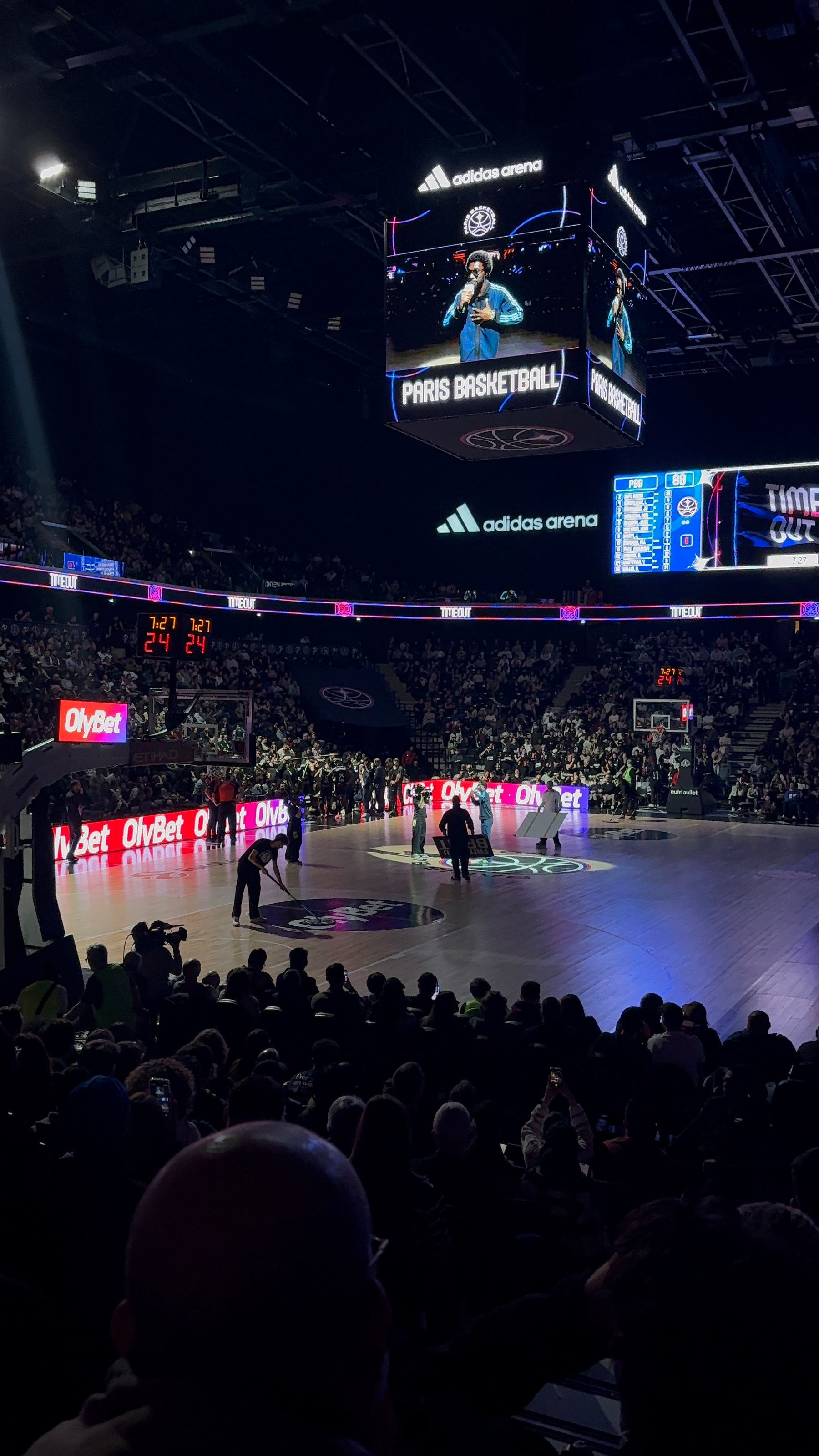 Fans in a sports arena watch a basketball game with a scoreboard displaying Paris Basketball and advertisements during a timeout.