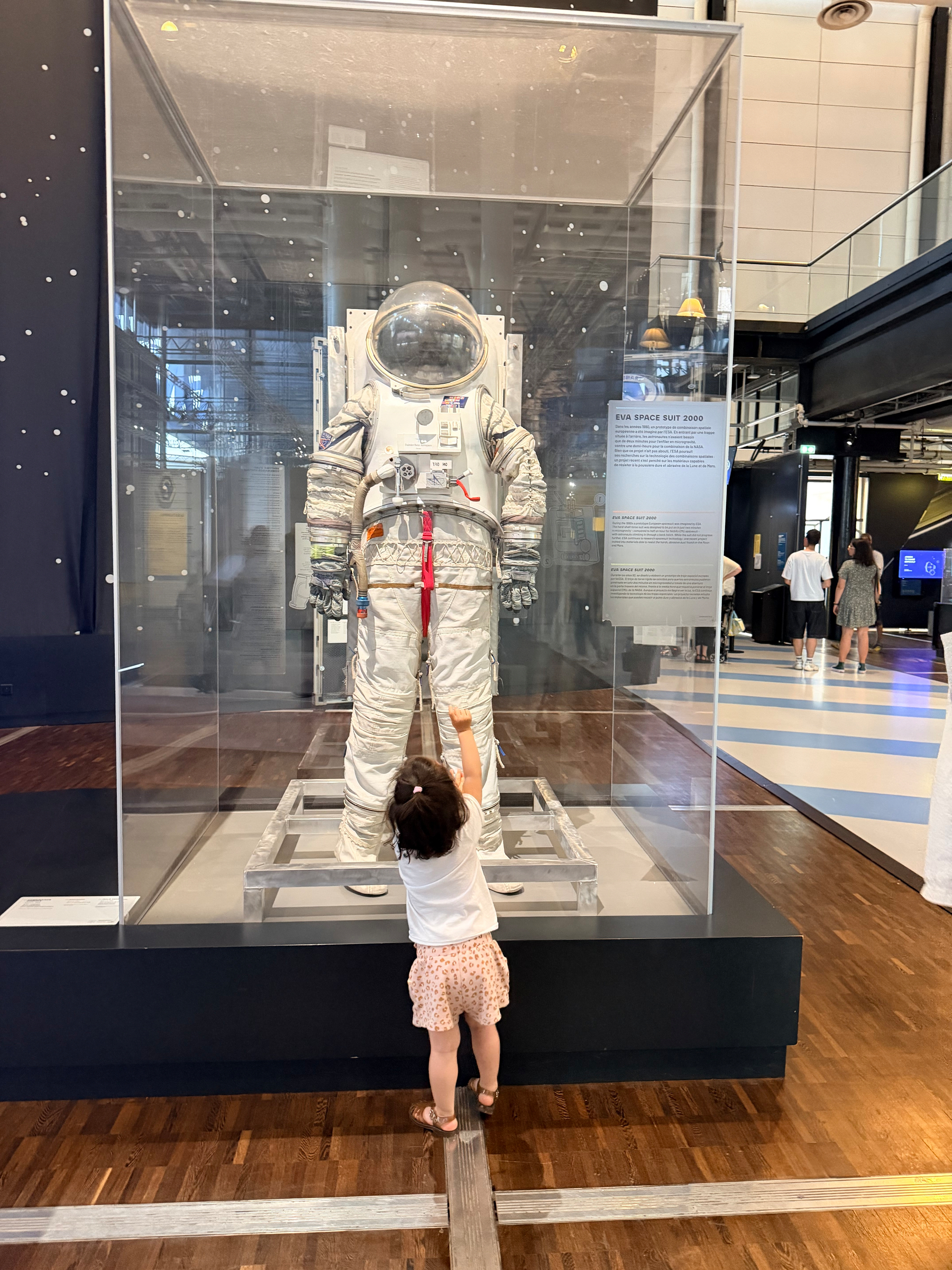 A small child reaches up towards a space suit displayed in a glass case at a museum.