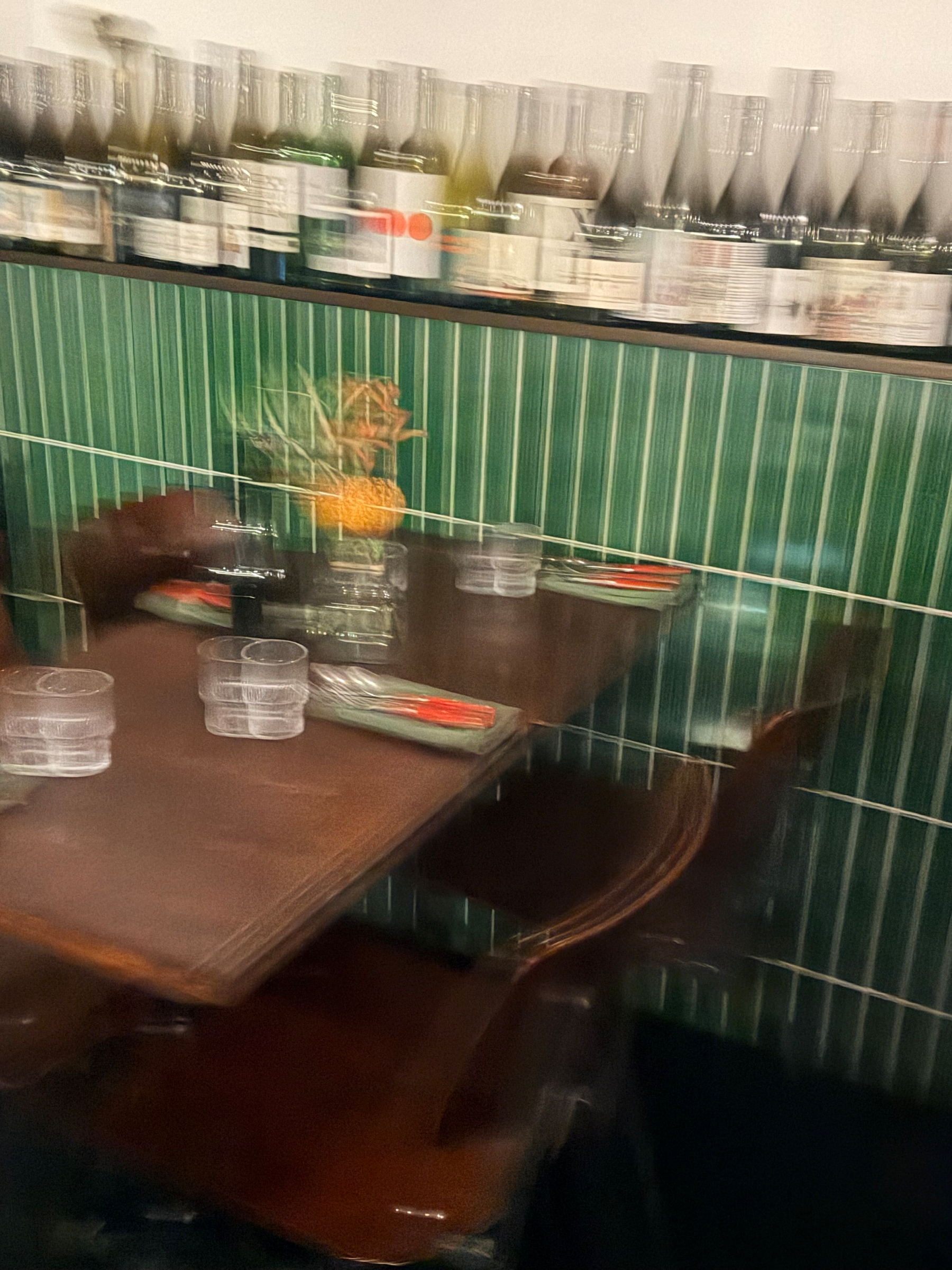 A dimly lit dining table is set with glasses and cutlery against a backdrop of green tiled walls and a shelf lined with bottles.