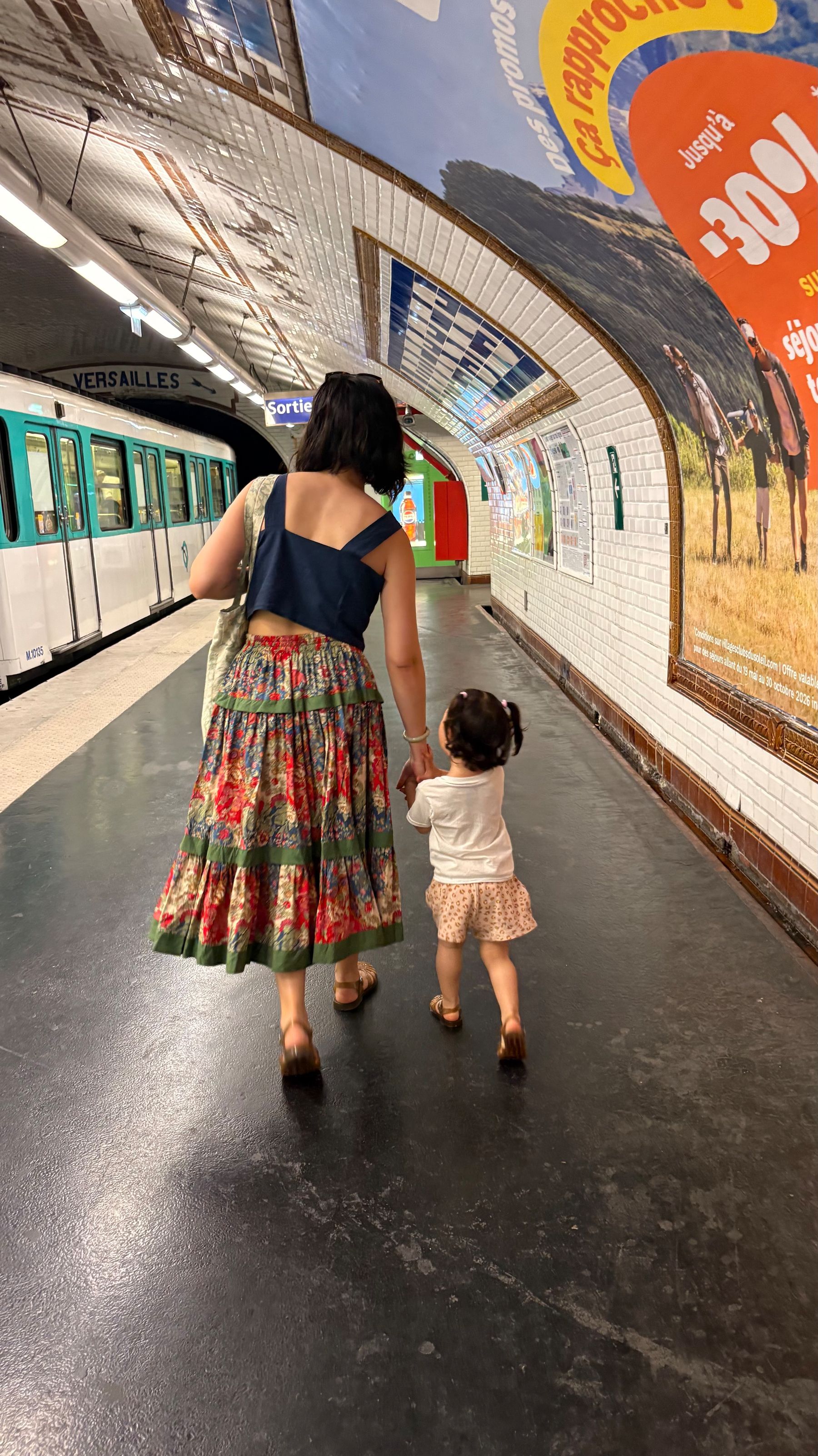 A woman and a toddler walk hand in hand down a tiled corridor of a metro station with a train on the left side.