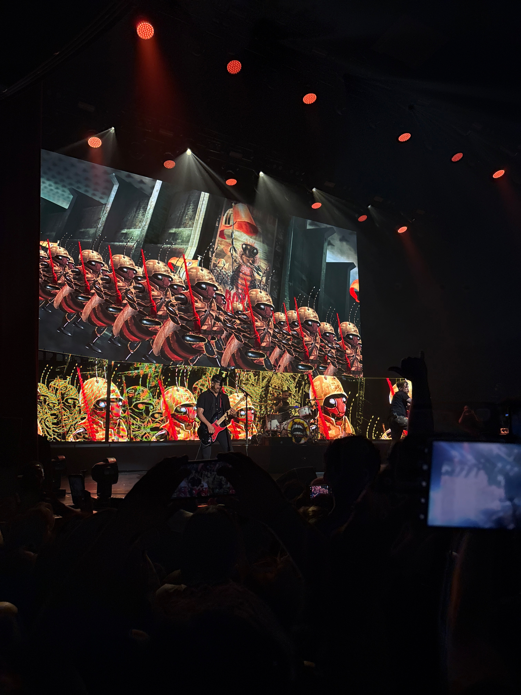 Auto-generated description: A live concert features musicians on stage with large screens displaying animated images of marching ants wearing hats and carrying batons.