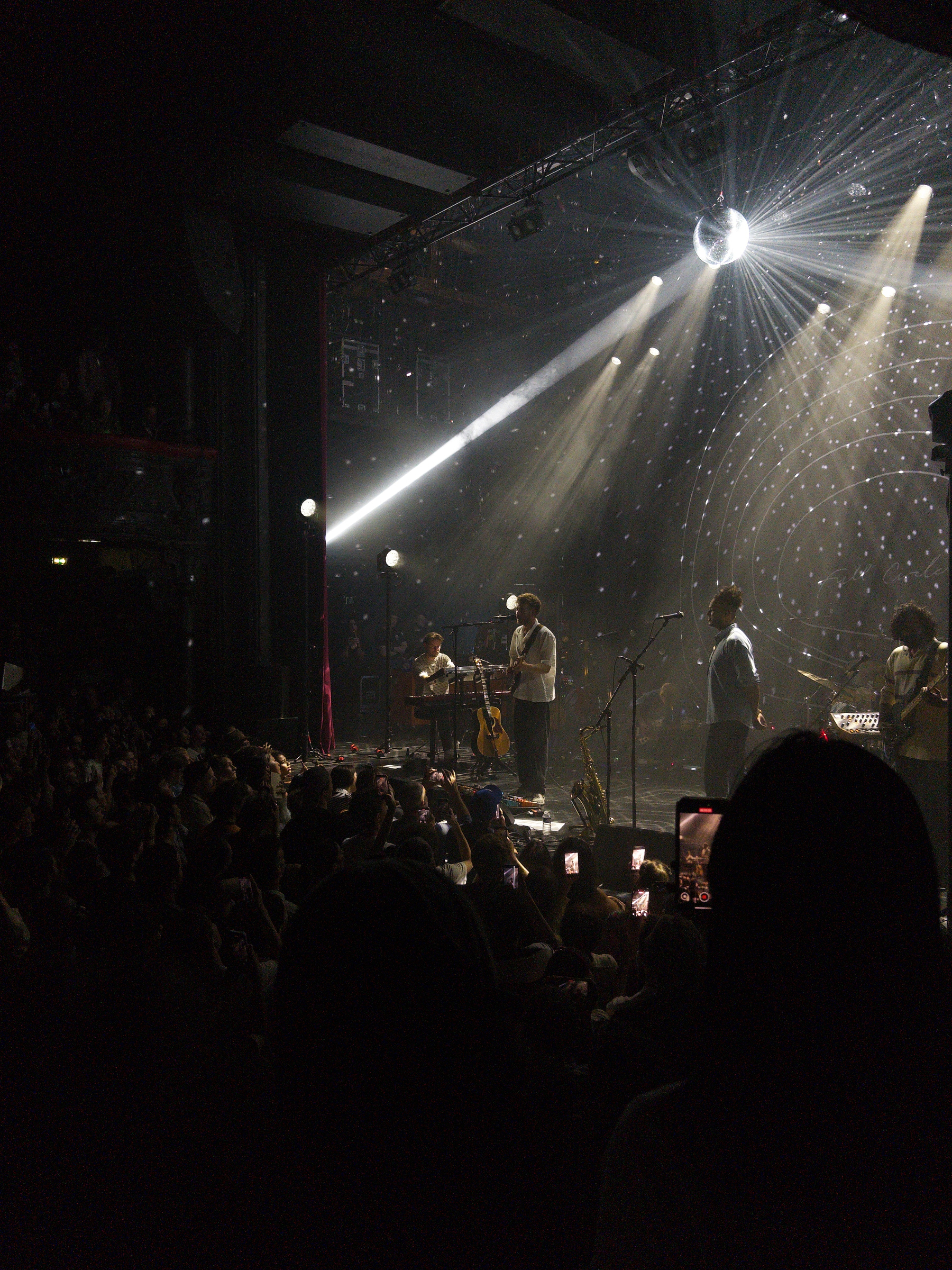 Auto-generated description: A live music performance is taking place on a stage with colorful lighting, surrounded by an engaged audience.