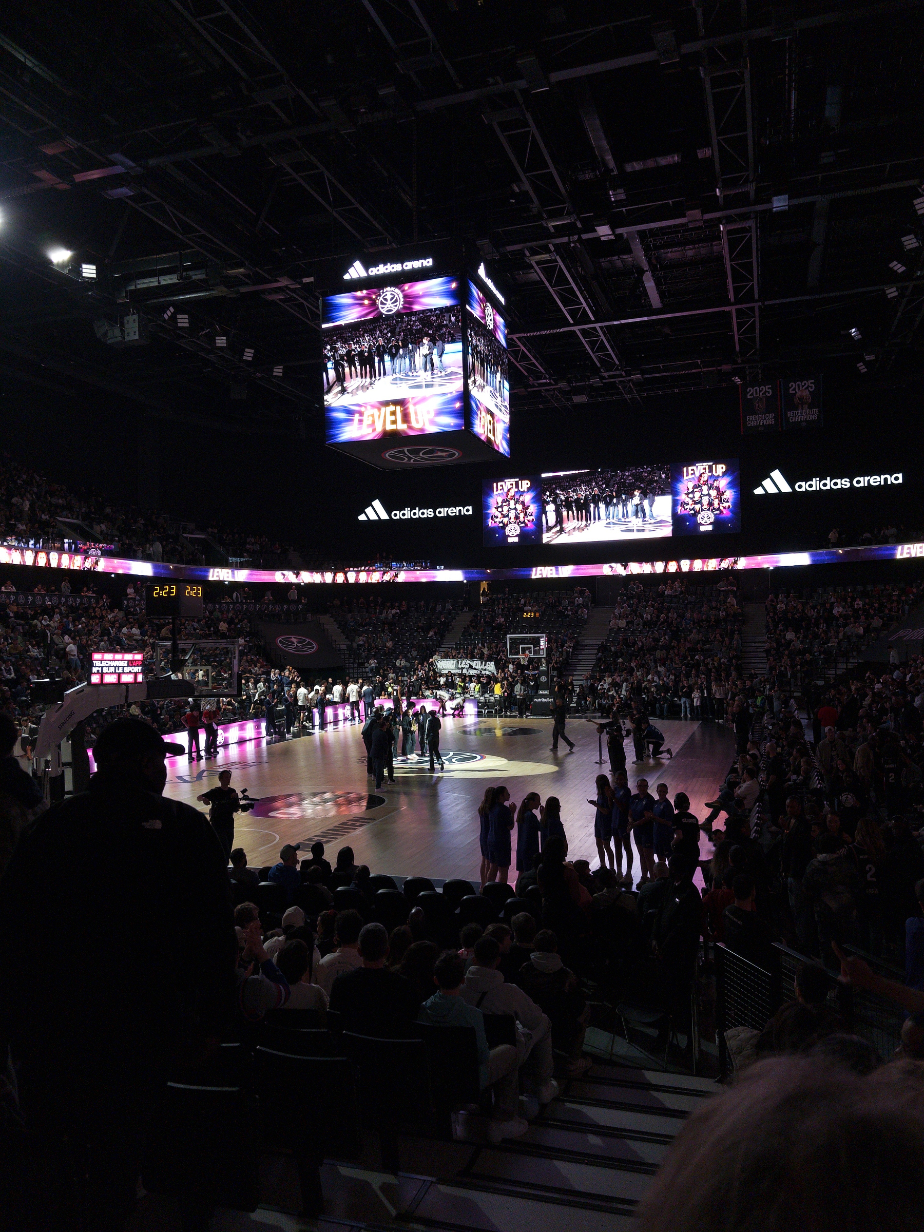 A basketball court inside a packed arena is illuminated with vibrant lights and large digital screens displaying visuals.