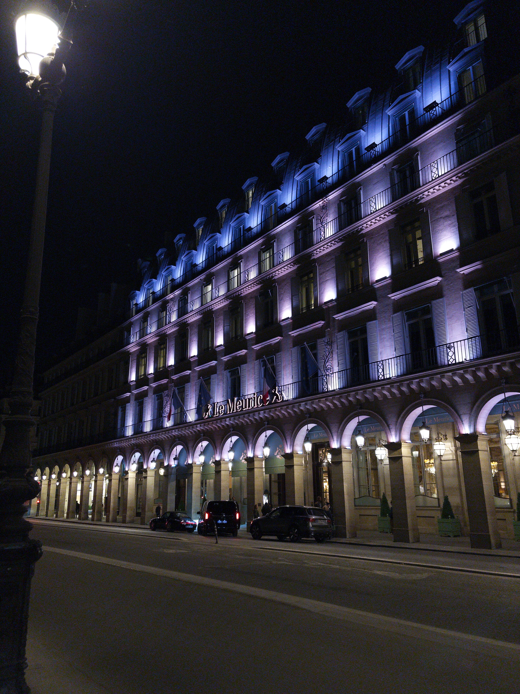 A grand building with arched windows and purple lighting is elegantly illuminated at night, with cars parked along the street and a lamp post in the foreground.