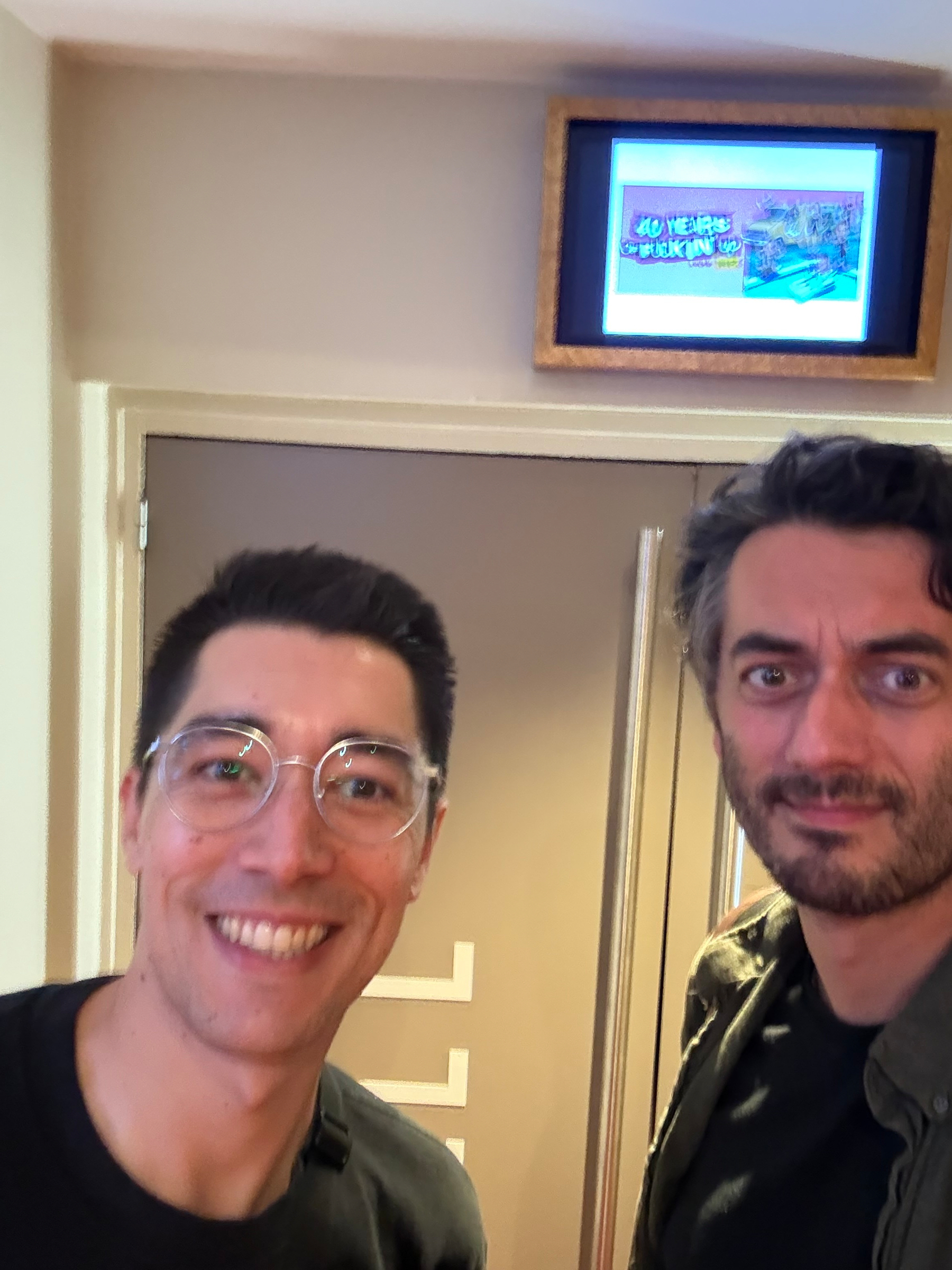 Two men are smiling and posing for a selfie in front of a door with a framed poster above it.