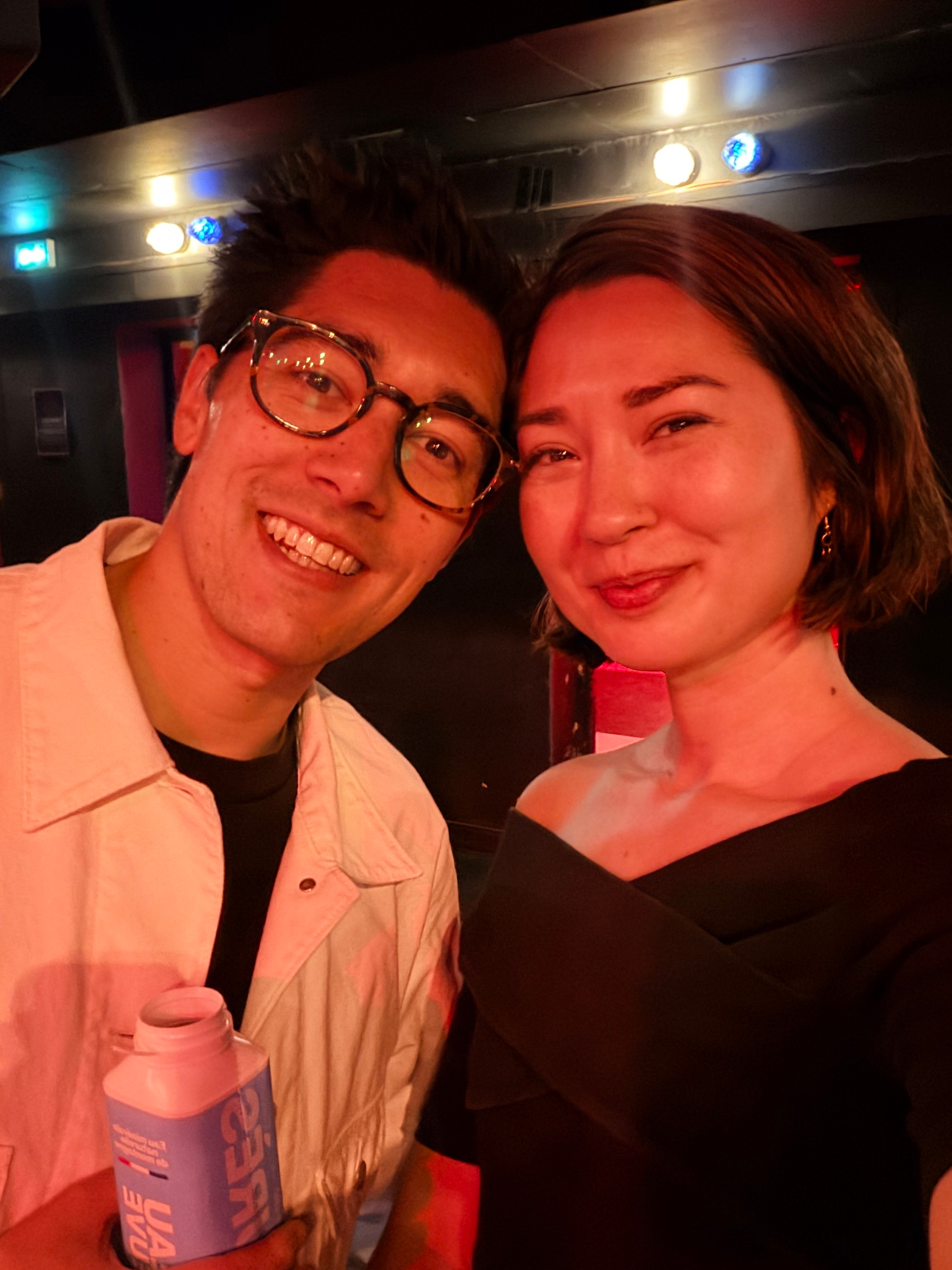 Auto-generated description: A smiling man holding a bottle and a woman in a dimly lit setting, possibly at an event.