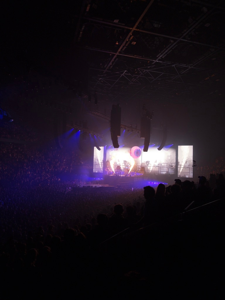 A large crowd is watching a live concert with vibrant stage lighting and visual effects in a dark indoor arena.