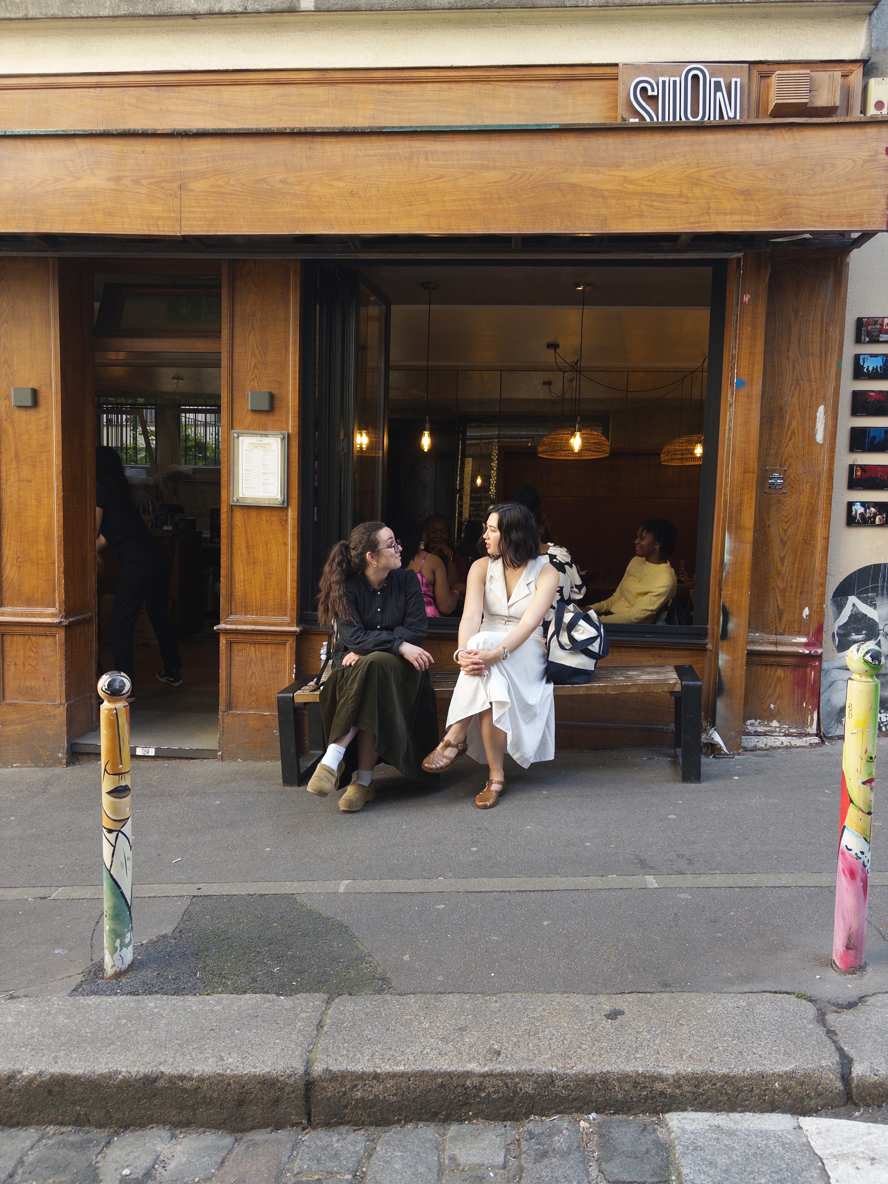 Auto-generated description: Two people are sitting and conversing outside a café with wooden exteriors and a partially open entrance.
