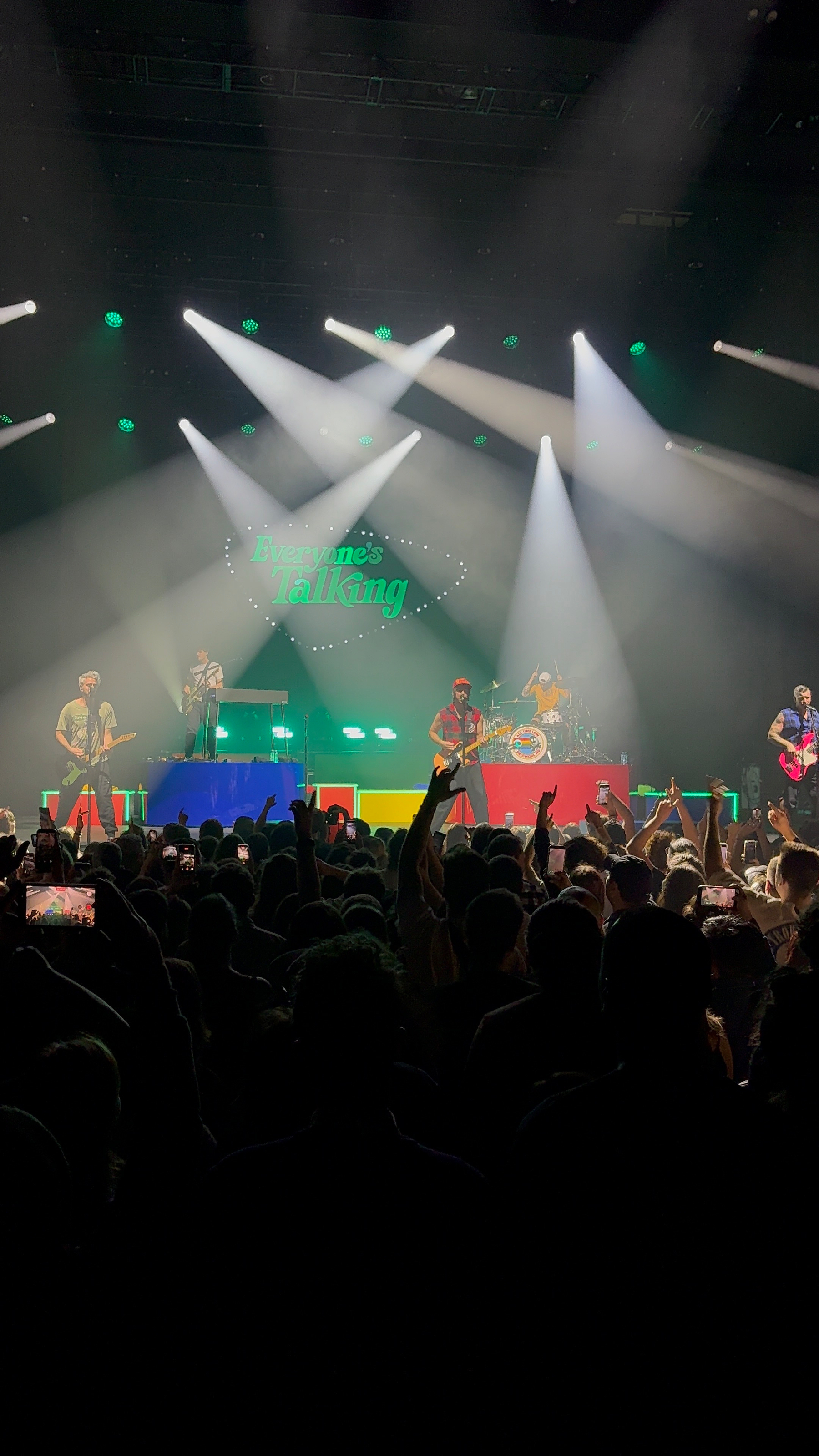A band performs energetically on a colorfully lit stage in front of a cheering crowd.