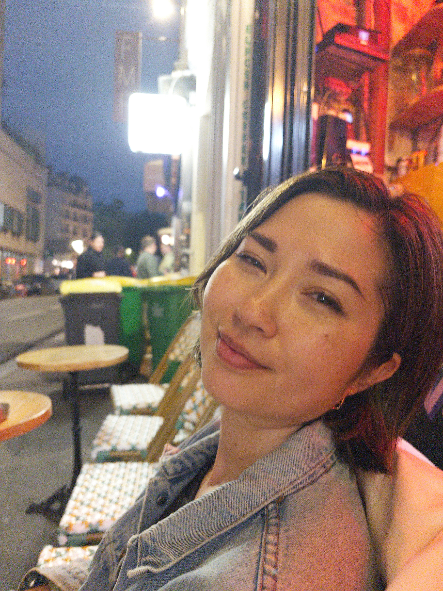 Auto-generated description: A person is sitting outside a café on a street with tables and chairs, smiling at the camera.