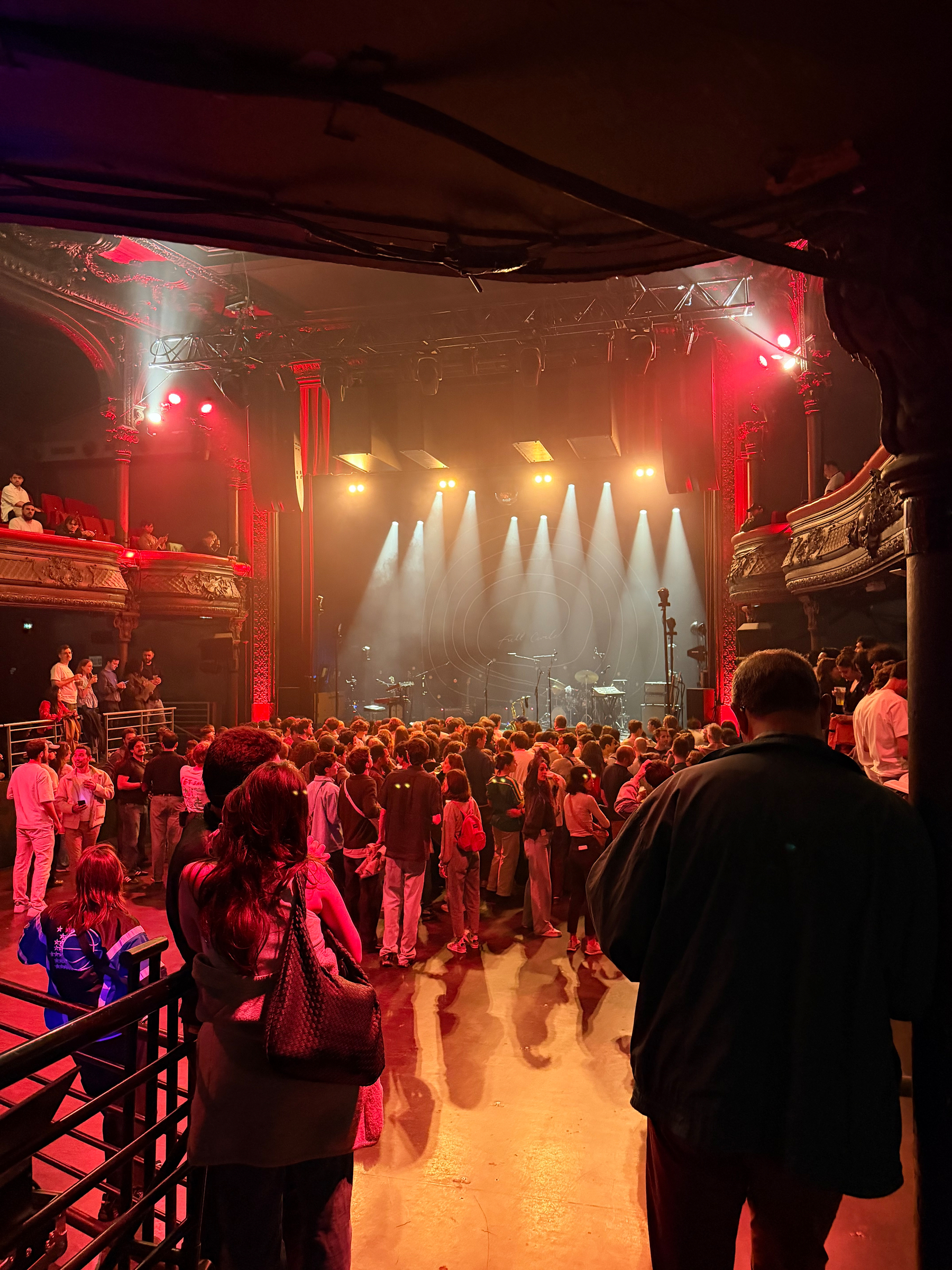 Auto-generated description: A crowd gathers in a dimly lit auditorium with dramatic lighting on the stage, preparing for a live performance.