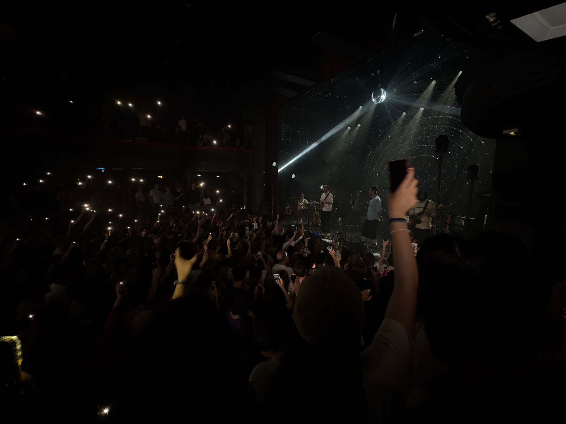 Auto-generated description: A lively concert scene with a large crowd holding up phones and lights, while a band performs on stage under a spotlight.