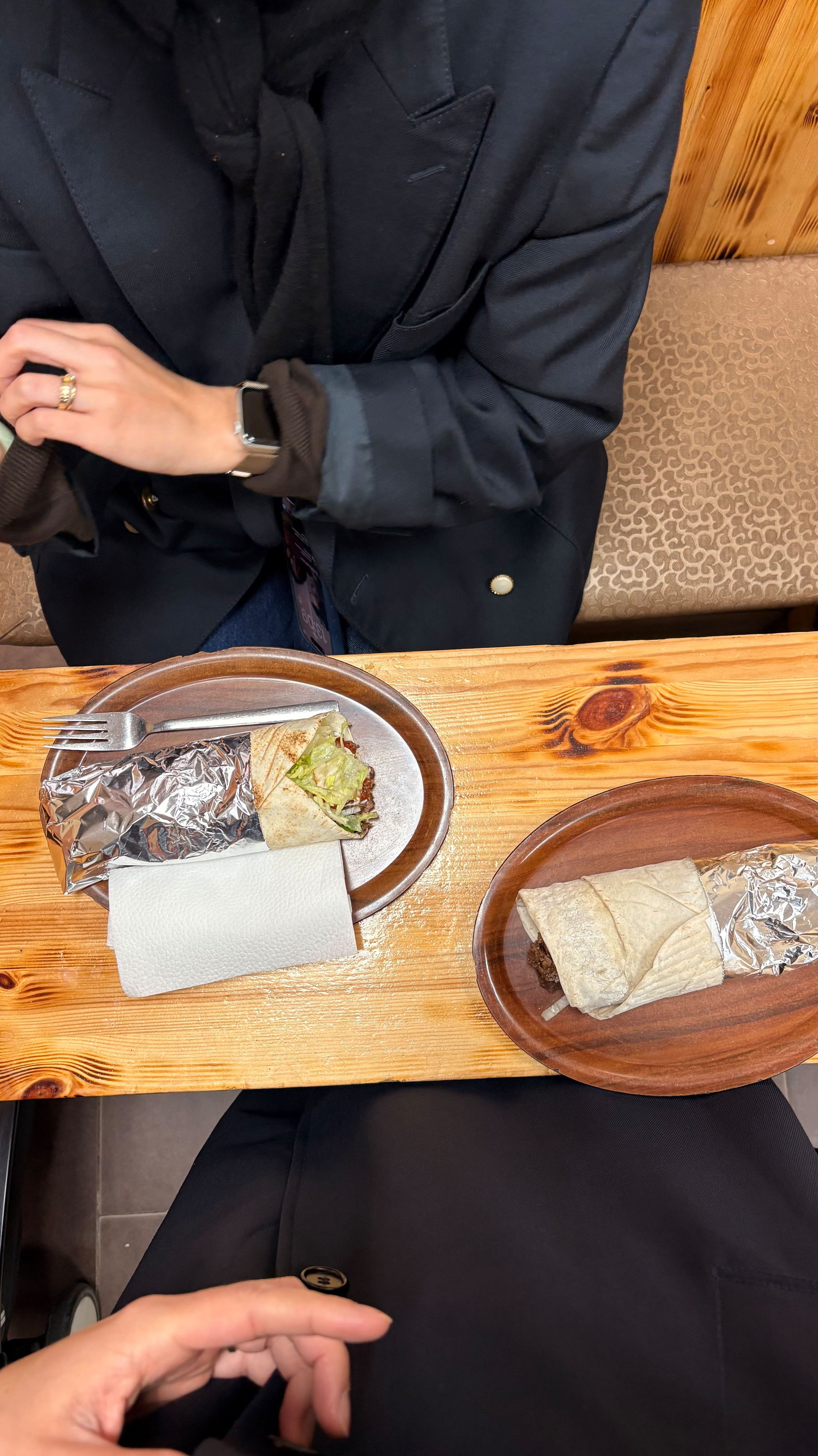 Auto-generated description: Two people are sitting at a wooden table with two burritos on plates, partially wrapped in foil.