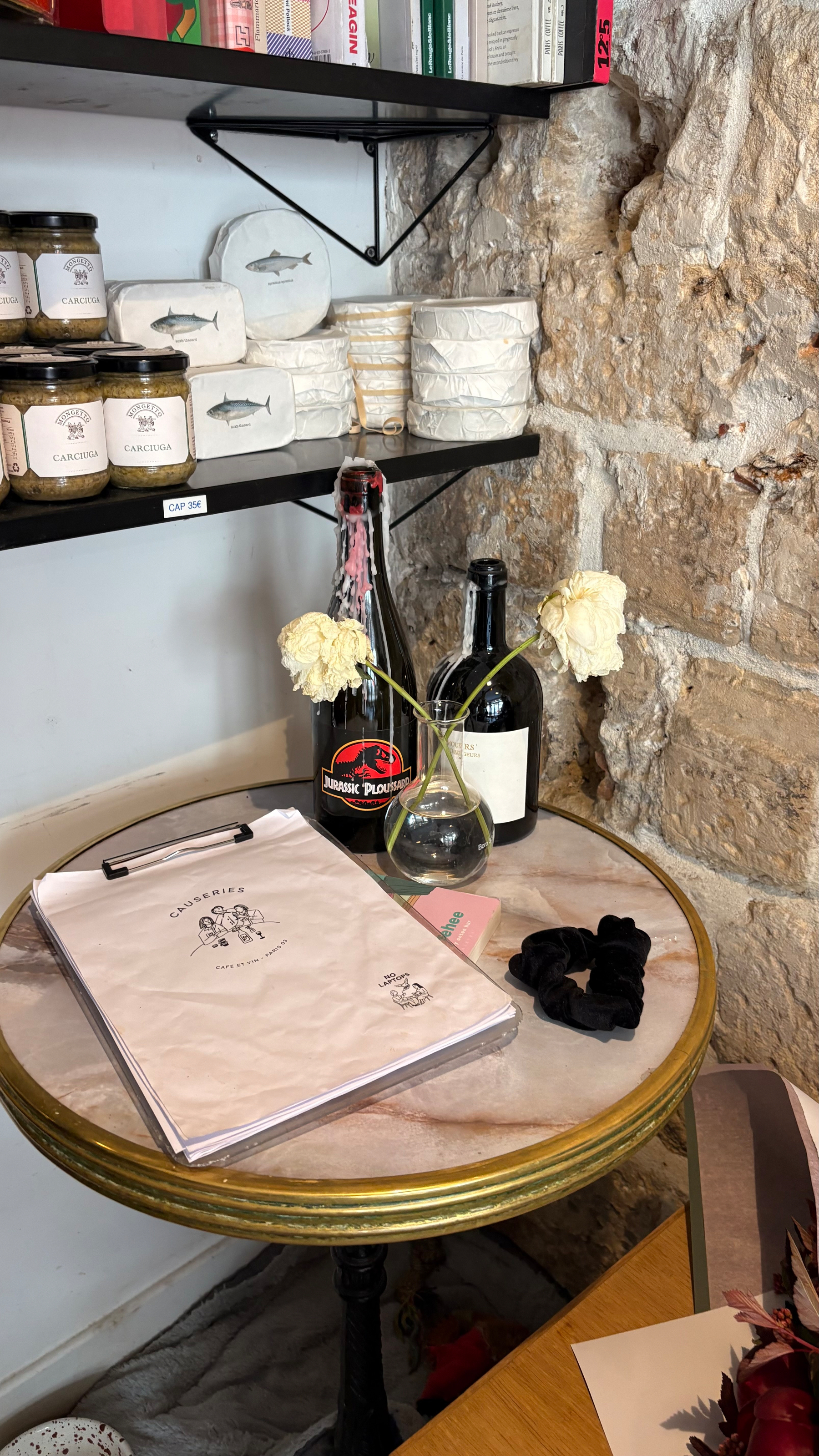 Auto-generated description: A small round table displays two white flowers in a vase, a clipboard, a black scrunchie, and two bottles, while shelves stocked with jars and wrapped items line the stone wall behind.
