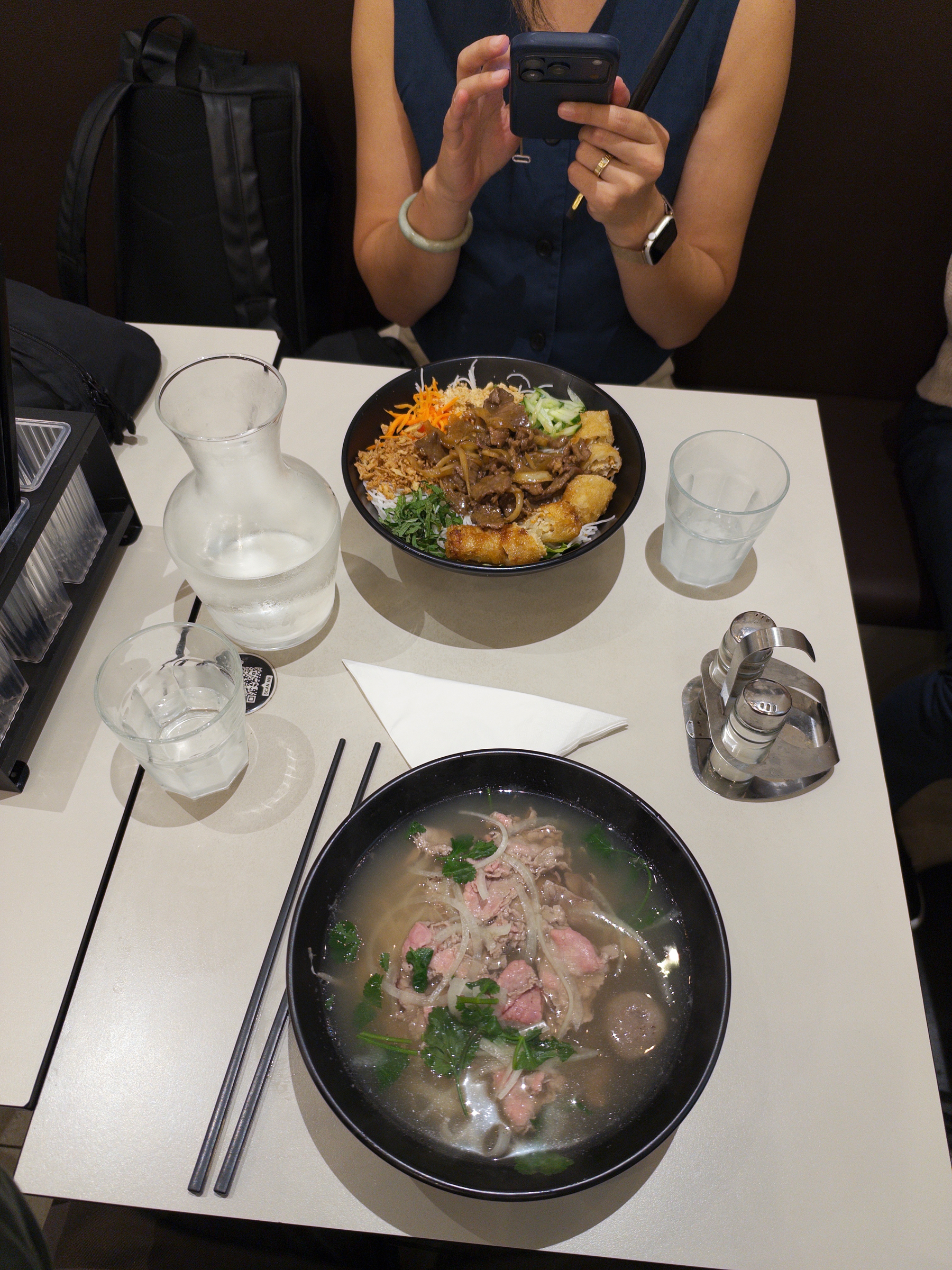 Auto-generated description: Two bowls of Asian-style dishes, one with soup and the other with various toppings, are set on a table along with glasses of water, chopsticks, a pitcher, and condiments, while a person sitting across is using their phone.