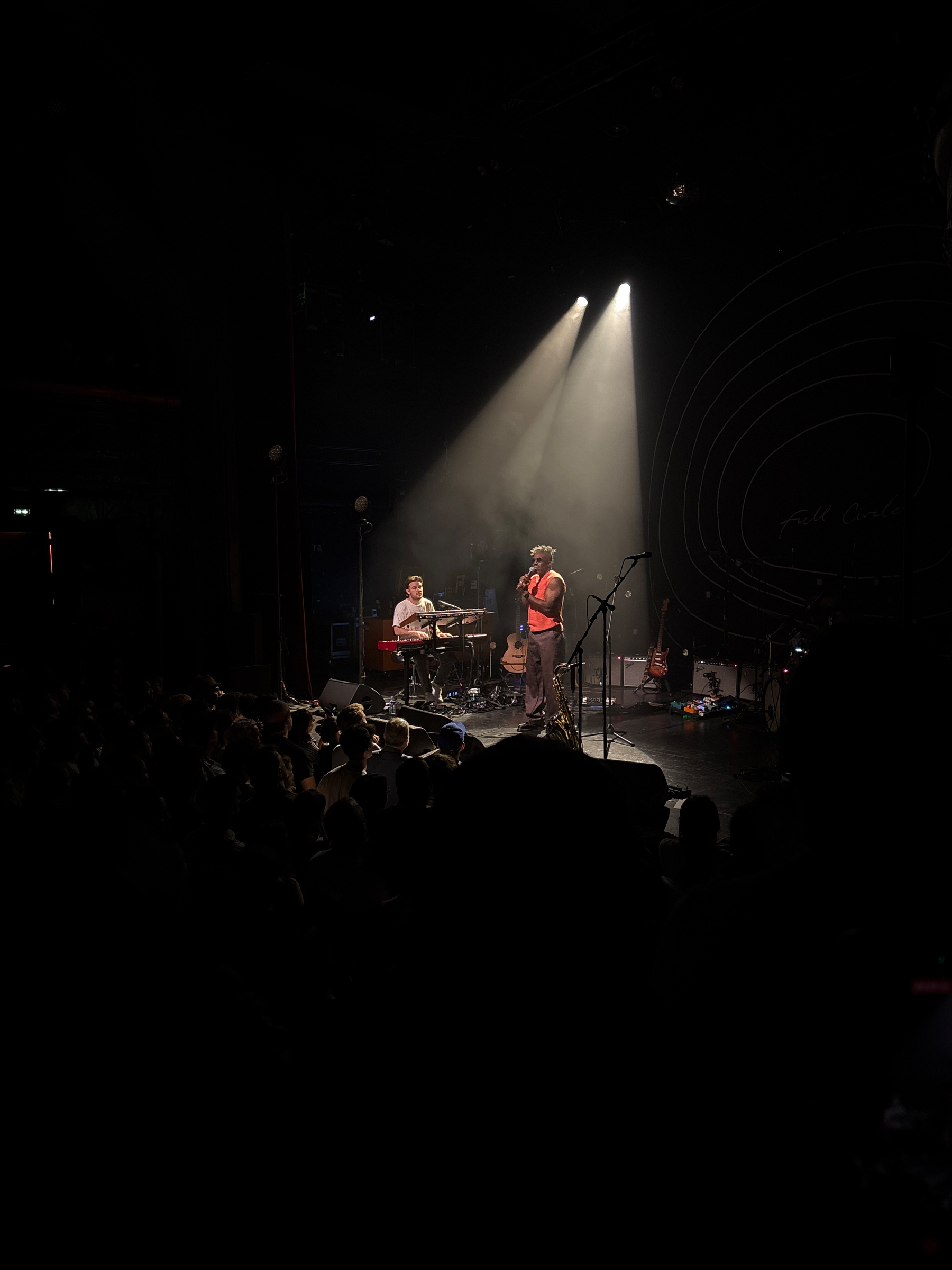 Auto-generated description: A live performance features two musicians on stage, one playing drums and the other singing into a microphone, under dramatic stage lighting.