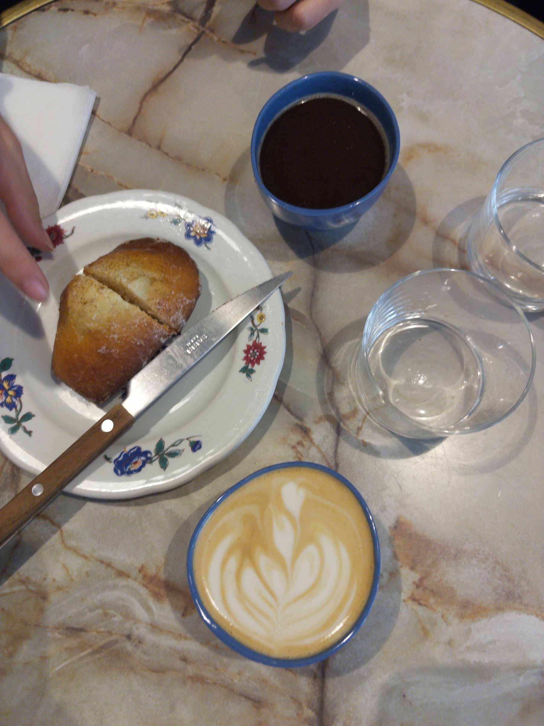 Auto-generated description: A cappuccino with latte art, a cup of hot chocolate, a pastry on a floral plate with a knife, and two glasses of water are arranged on a marble table.