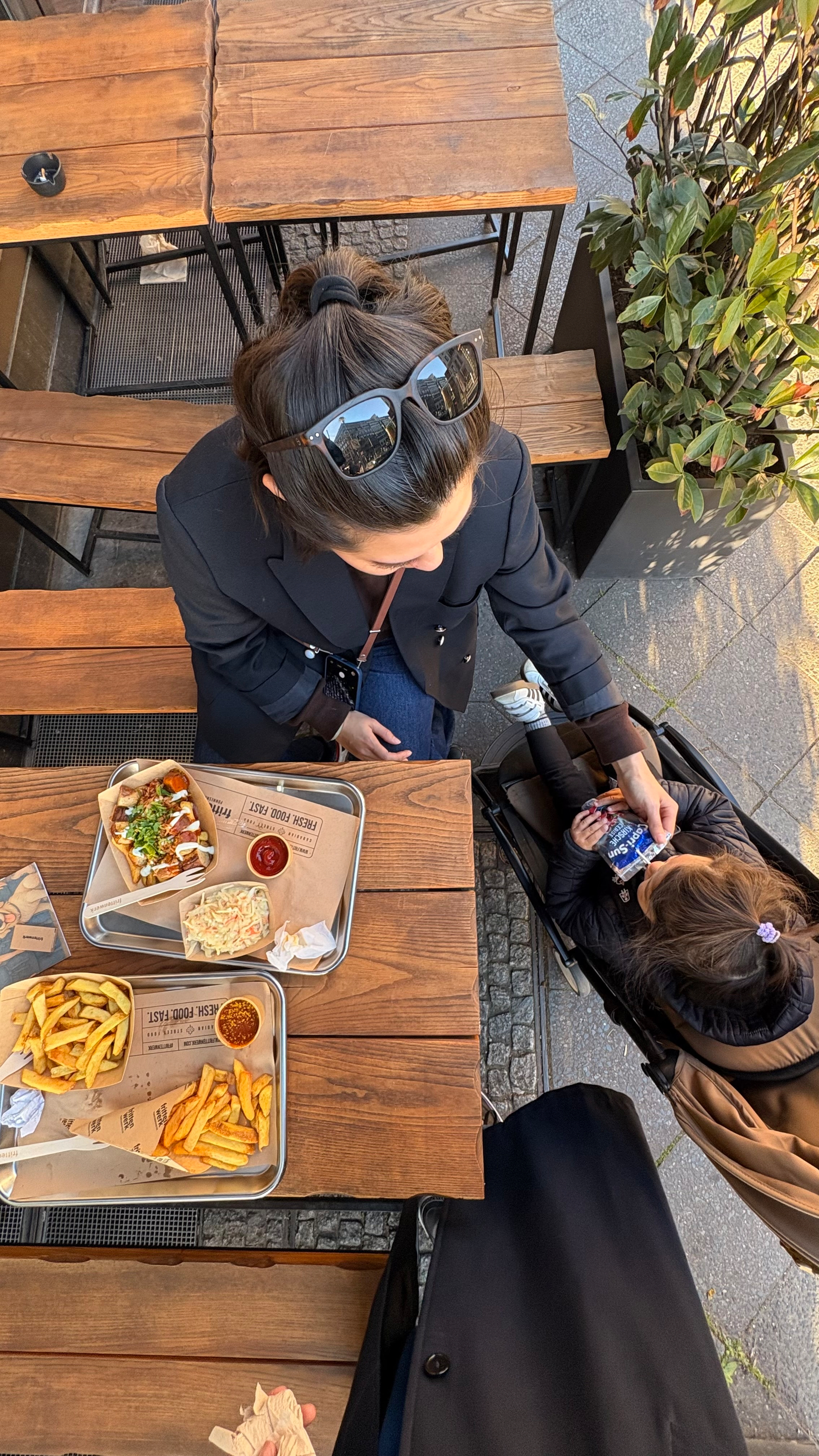 Auto-generated description: A woman is sitting at an outdoor table with food while tending to a child in a stroller next to her.