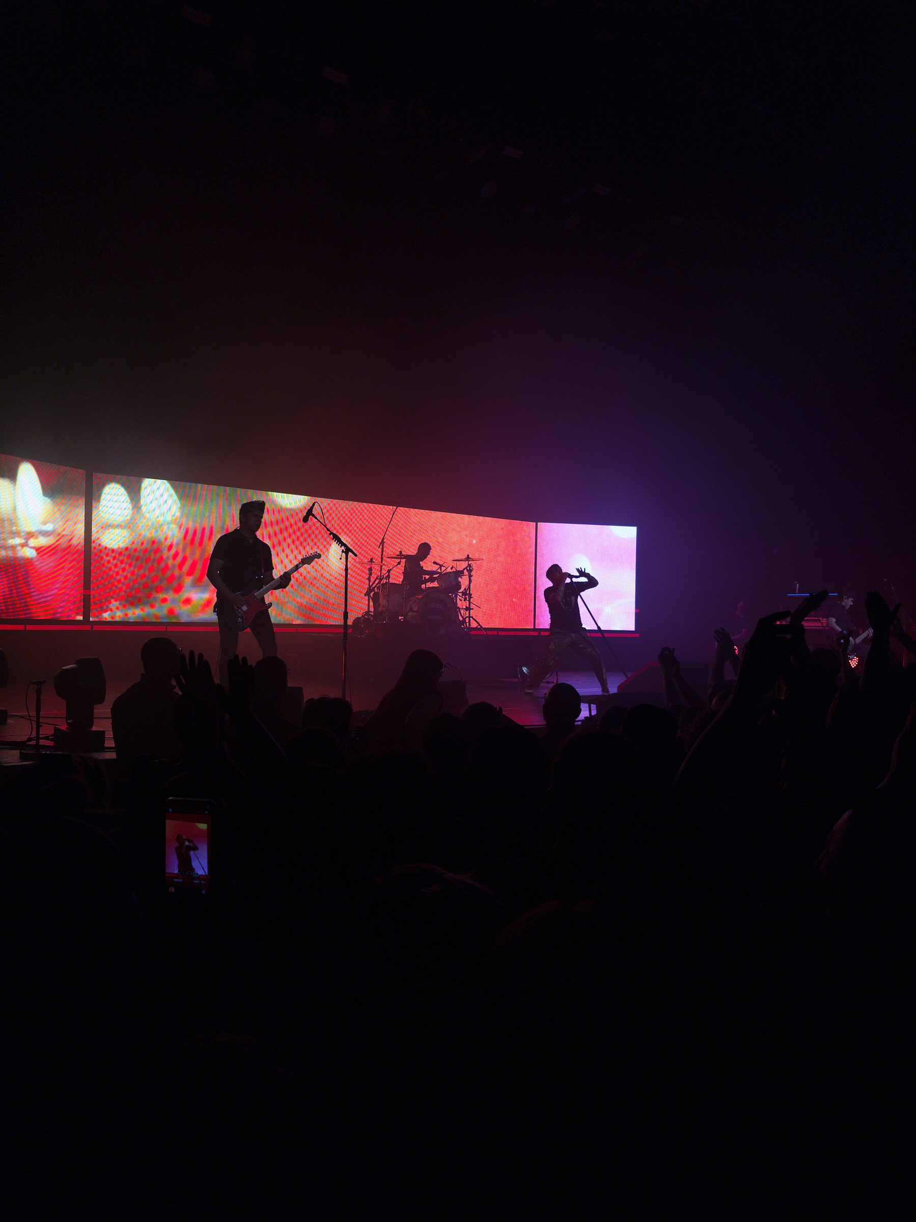 Auto-generated description: A live band performs on stage with bright, colorful screens behind them while an audience watches in the foreground.