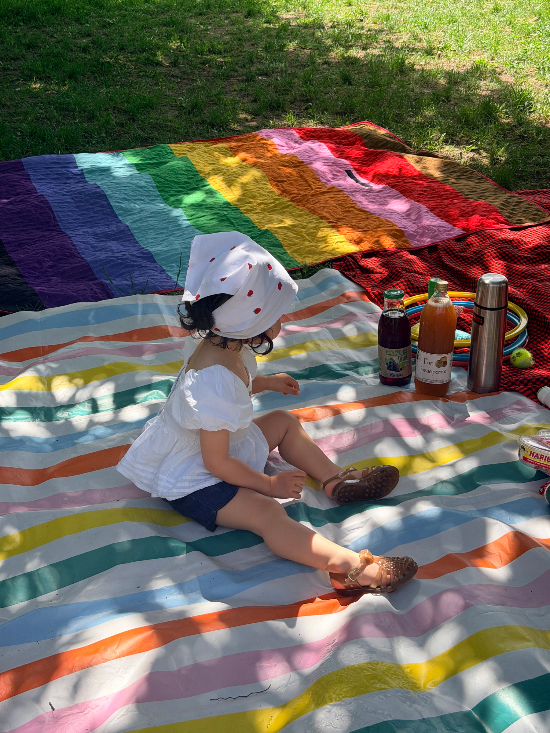 A child wearing a white hat is sitting on a colorful striped blanket, surrounded by drinks and snacks on the grass.