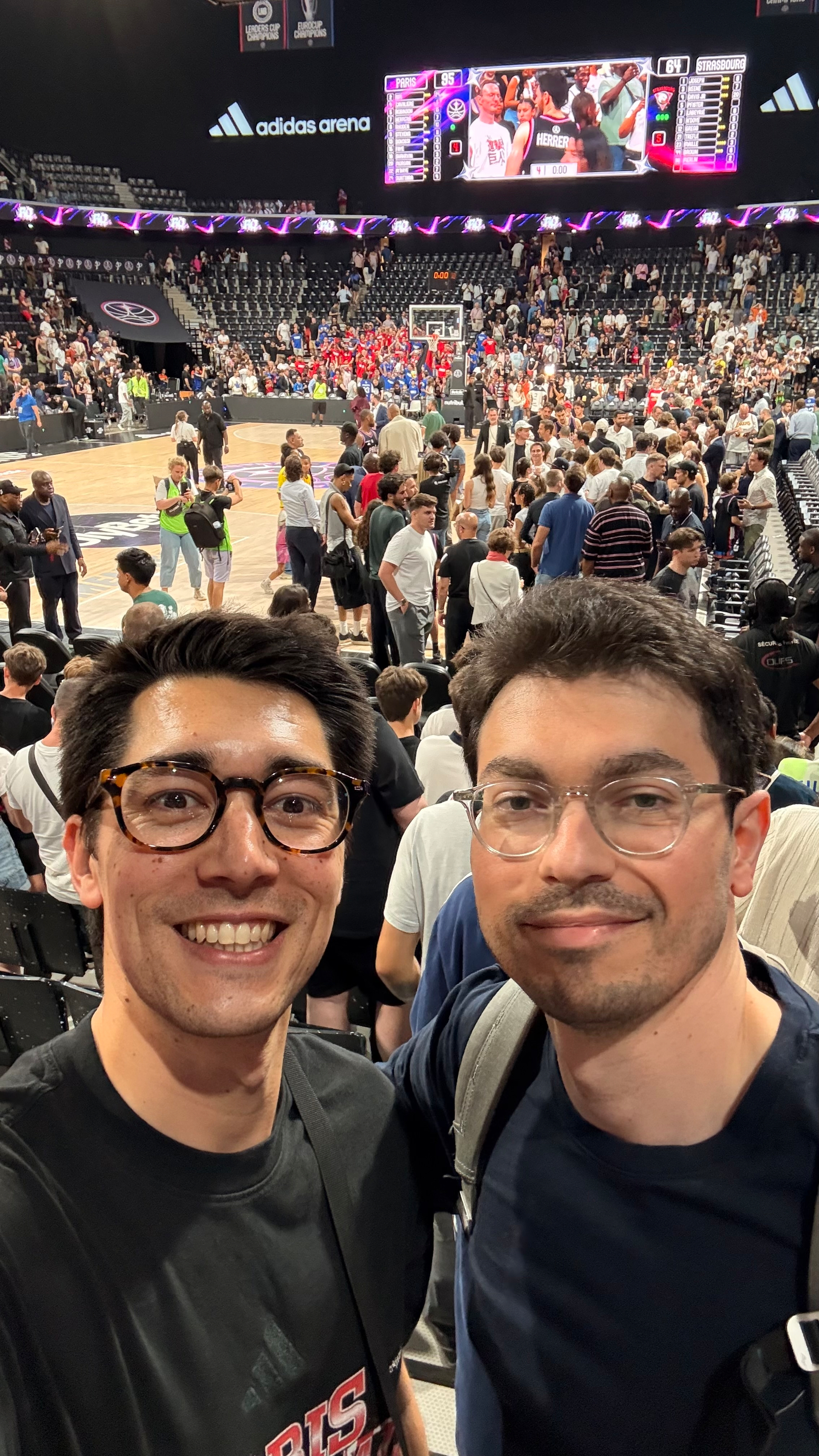 Two people take a selfie in a crowded basketball arena with a game taking place in the background.