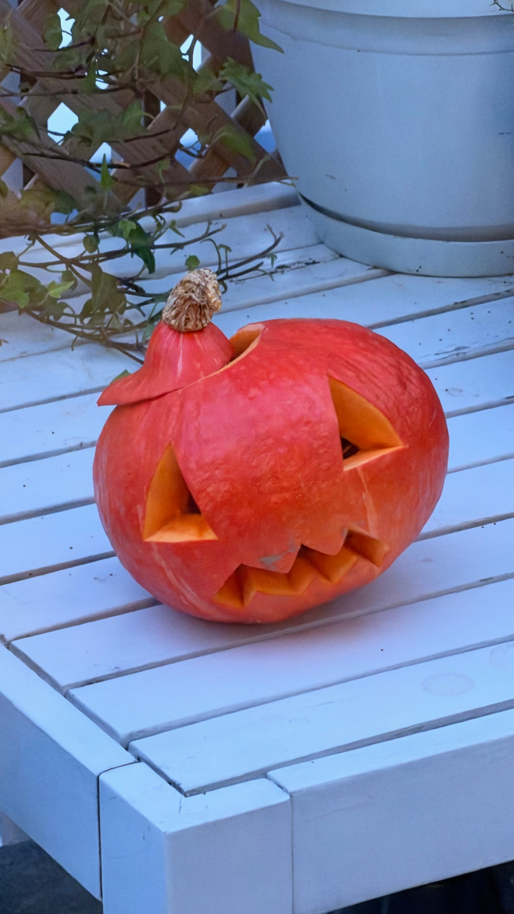 A carved pumpkin with triangular eyes and a jagged mouth is placed on a white wooden table near a plant.