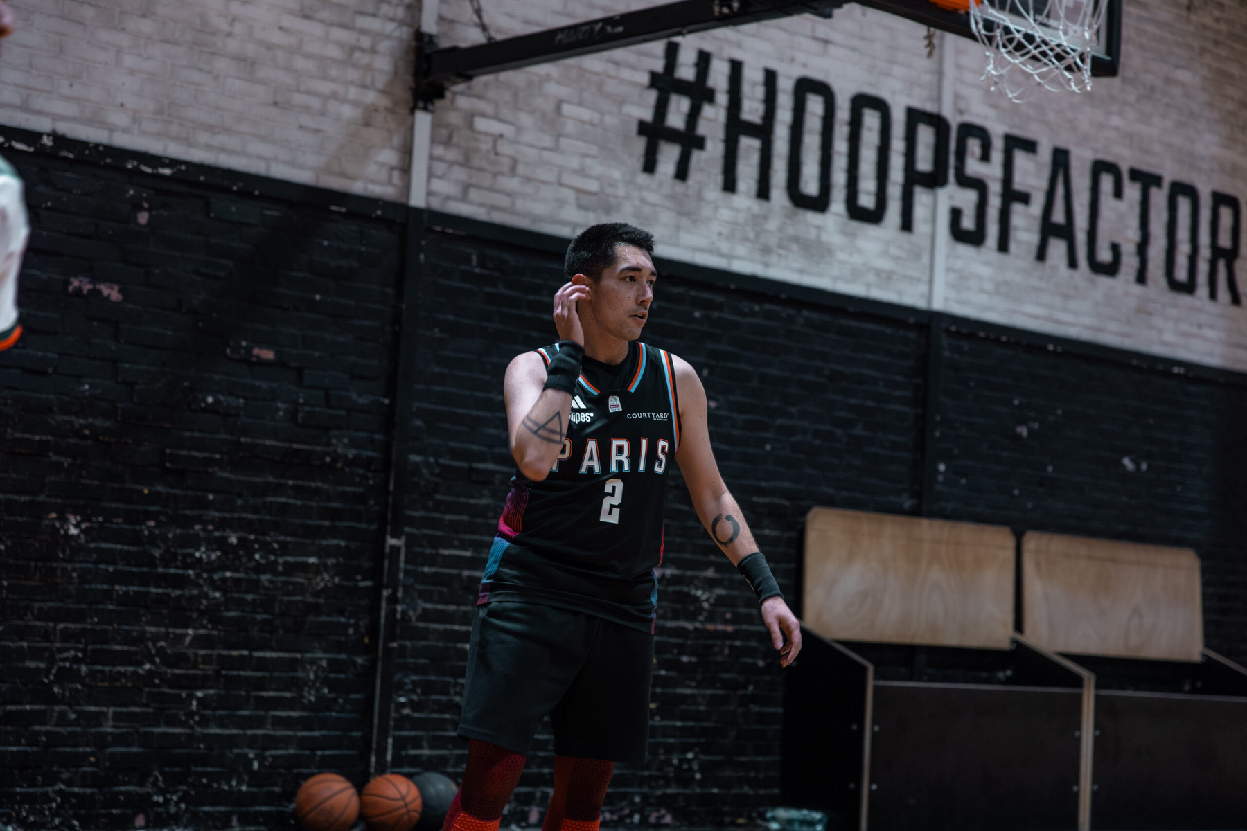 A basketball player wearing a jersey with the number 2 stands on an indoor court with the hashtag #HOOPSFACTORY on the wall behind him.