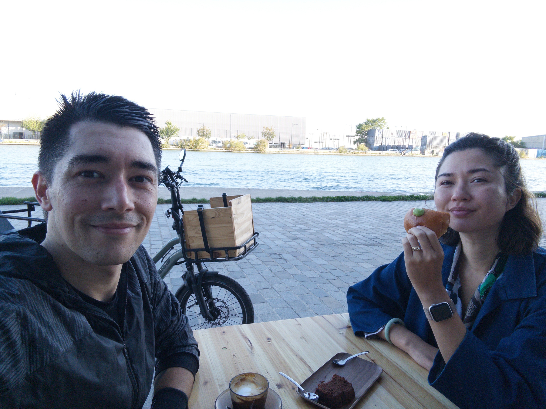 A man and a woman are sitting at an outdoor table near the water, with the woman eating a pastry and the man smiling at the camera.