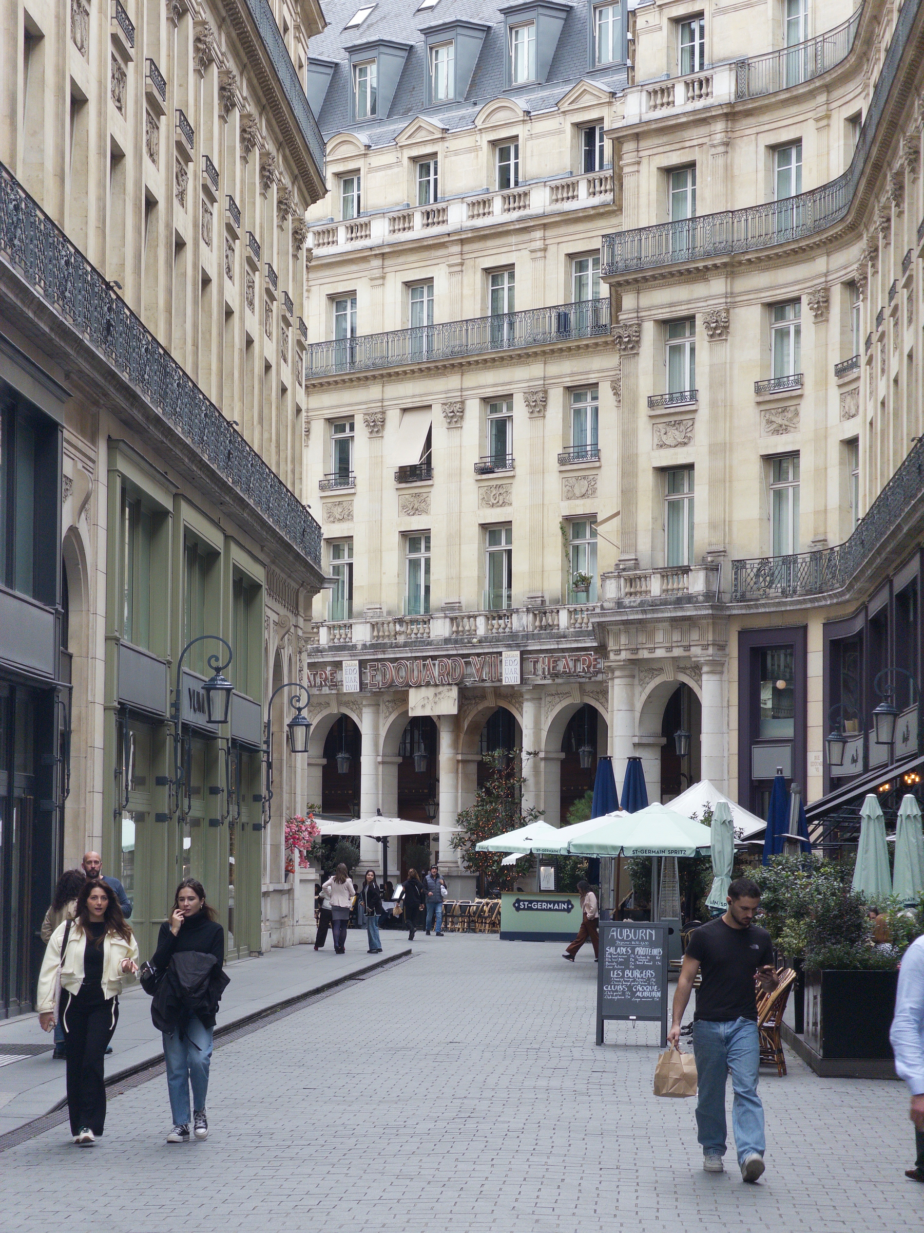 Auto-generated description: A European-style street scene features people walking and dining along a cobblestone pathway lined with historic buildings and outdoor café seating.