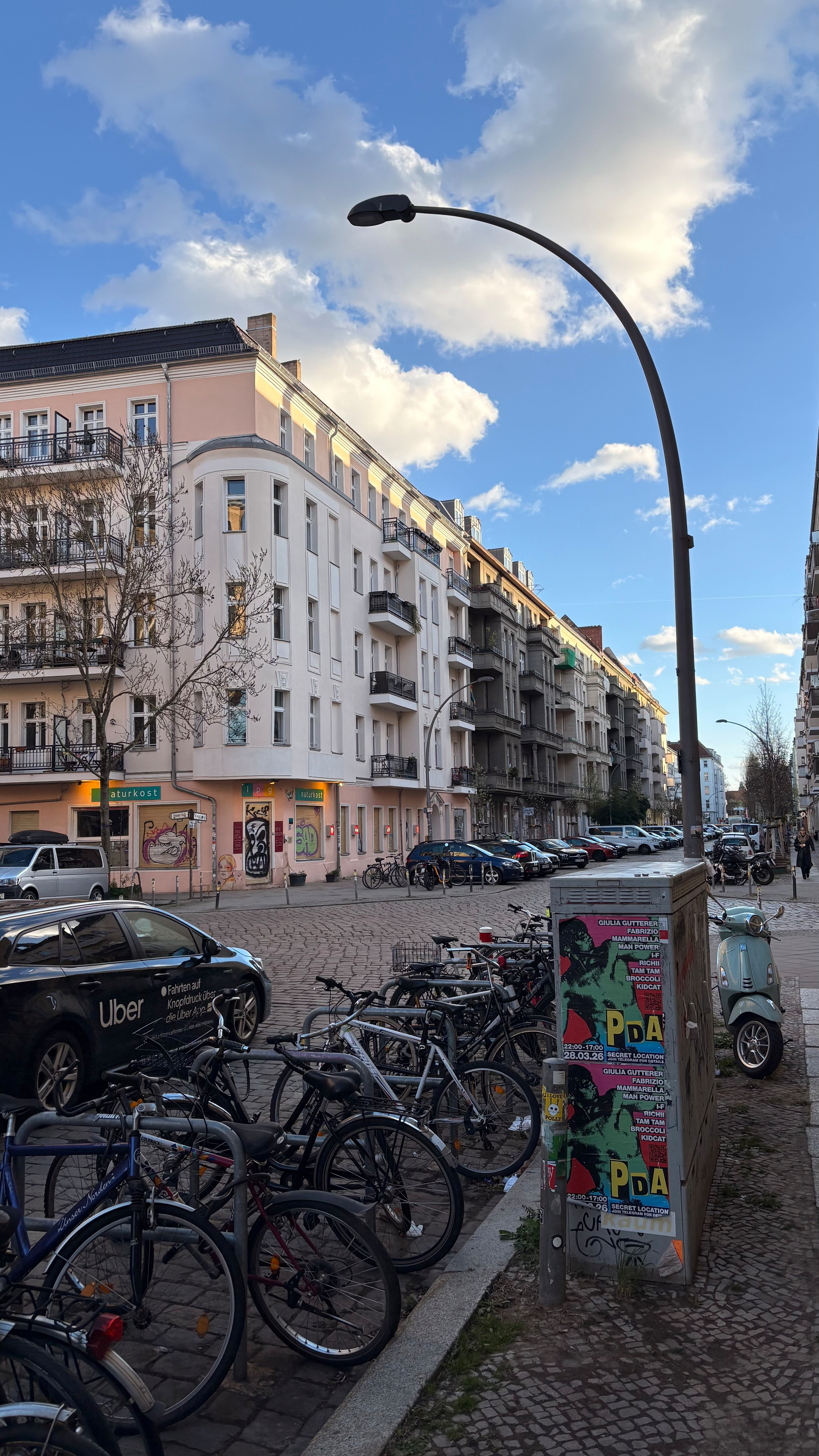 Auto-generated description: A street scene features a row of bicycles parked along a cobblestone sidewalk, with residential buildings and a street lamp against a partially cloudy sky.