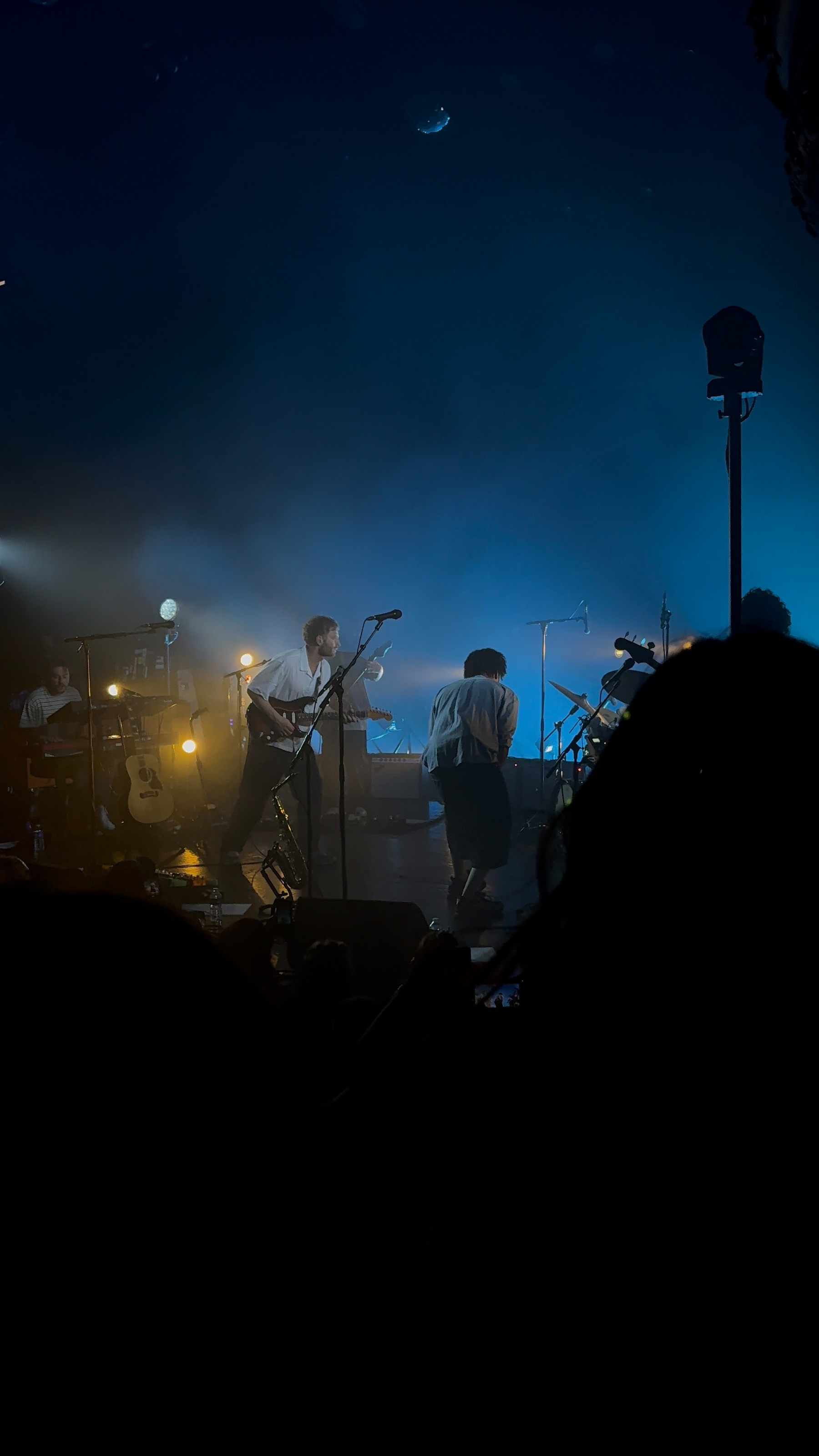 Auto-generated description: A band performs energetically on stage under moody blue and white lighting, surrounded by musical instruments.