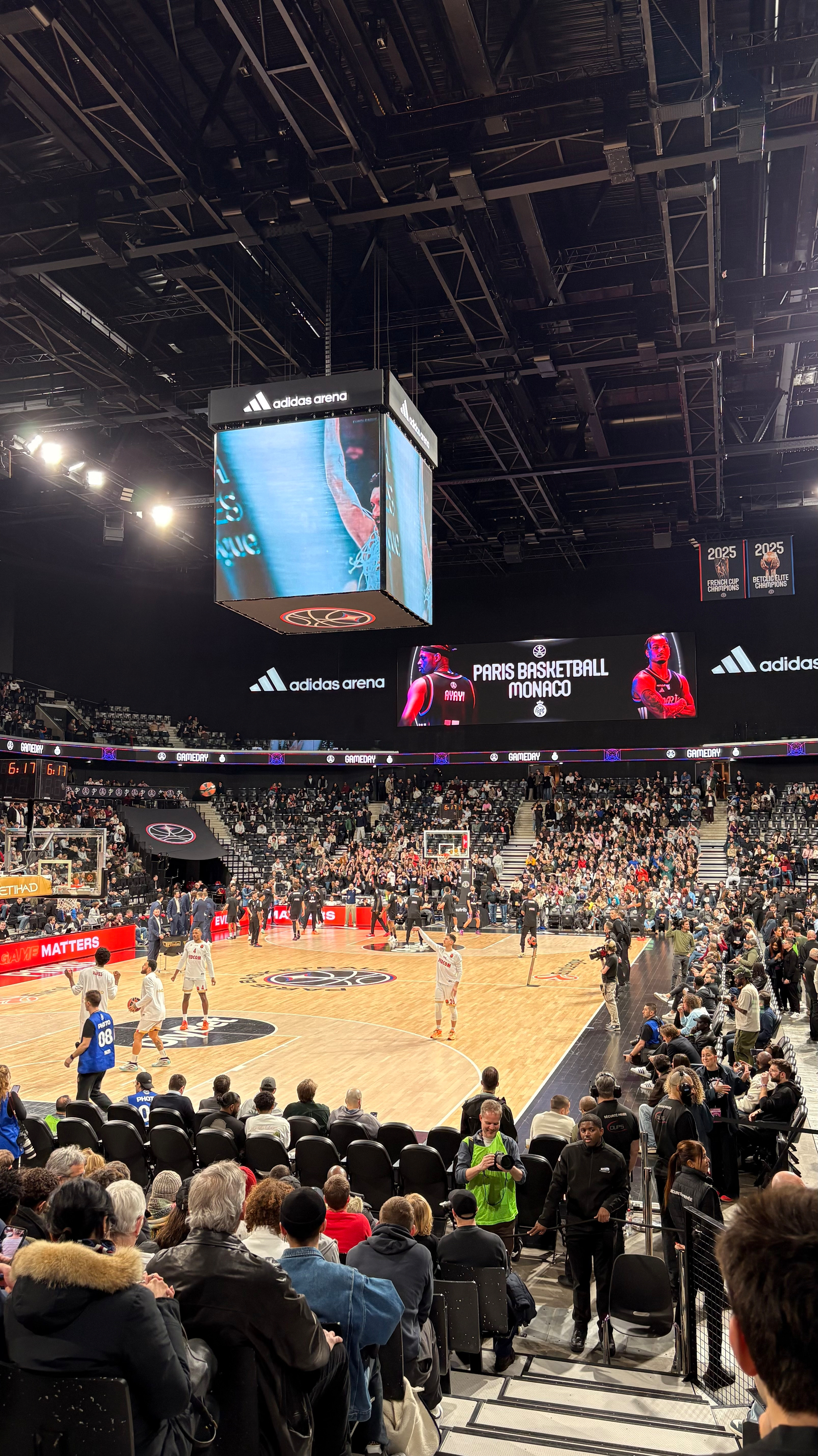 A basketball game is taking place in an arena with spectators watching and a large screen overhead displaying game information.